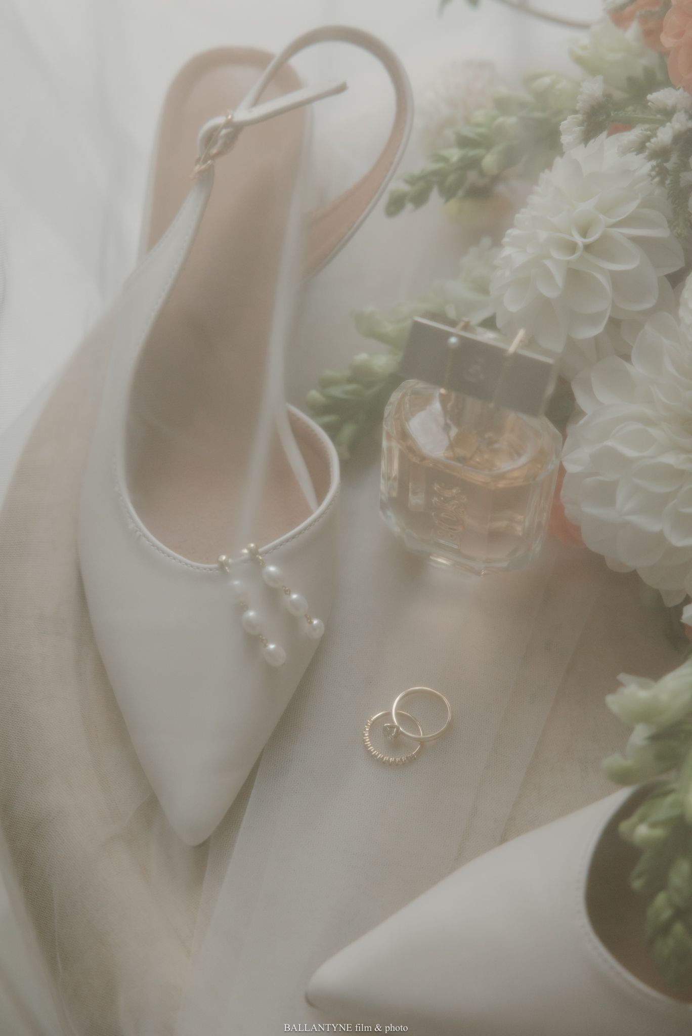 Elegant bridal flat shoes with pearl details, gold wedding rings, perfume bottle, and white floral bouquet arranged beautifully.