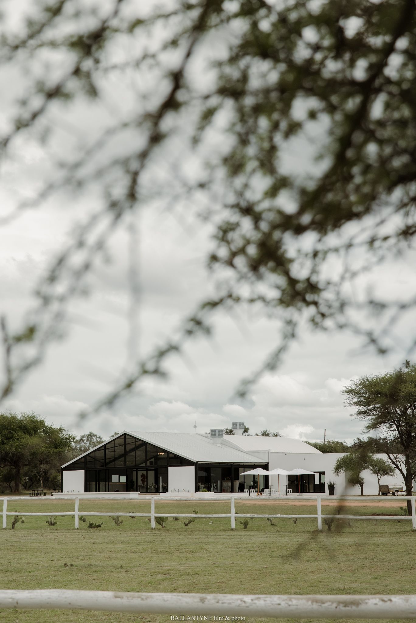 White barn wedding venue framed by tree branches on a green farm under a cloudy sky.