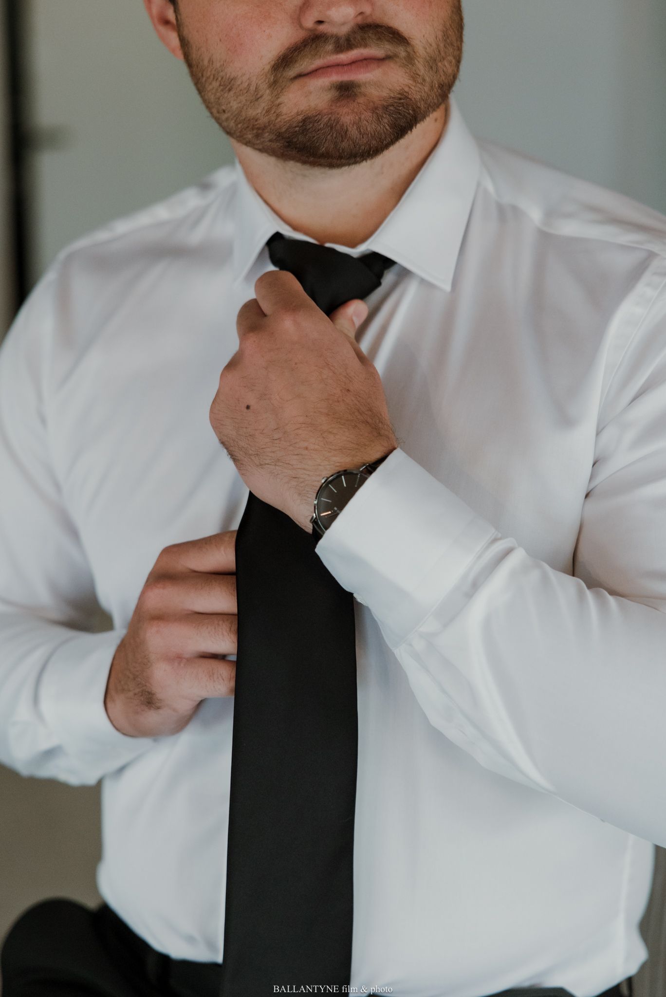 Man in white dress shirt adjusting black necktie, wearing a watch, getting dressed for formal occasion.