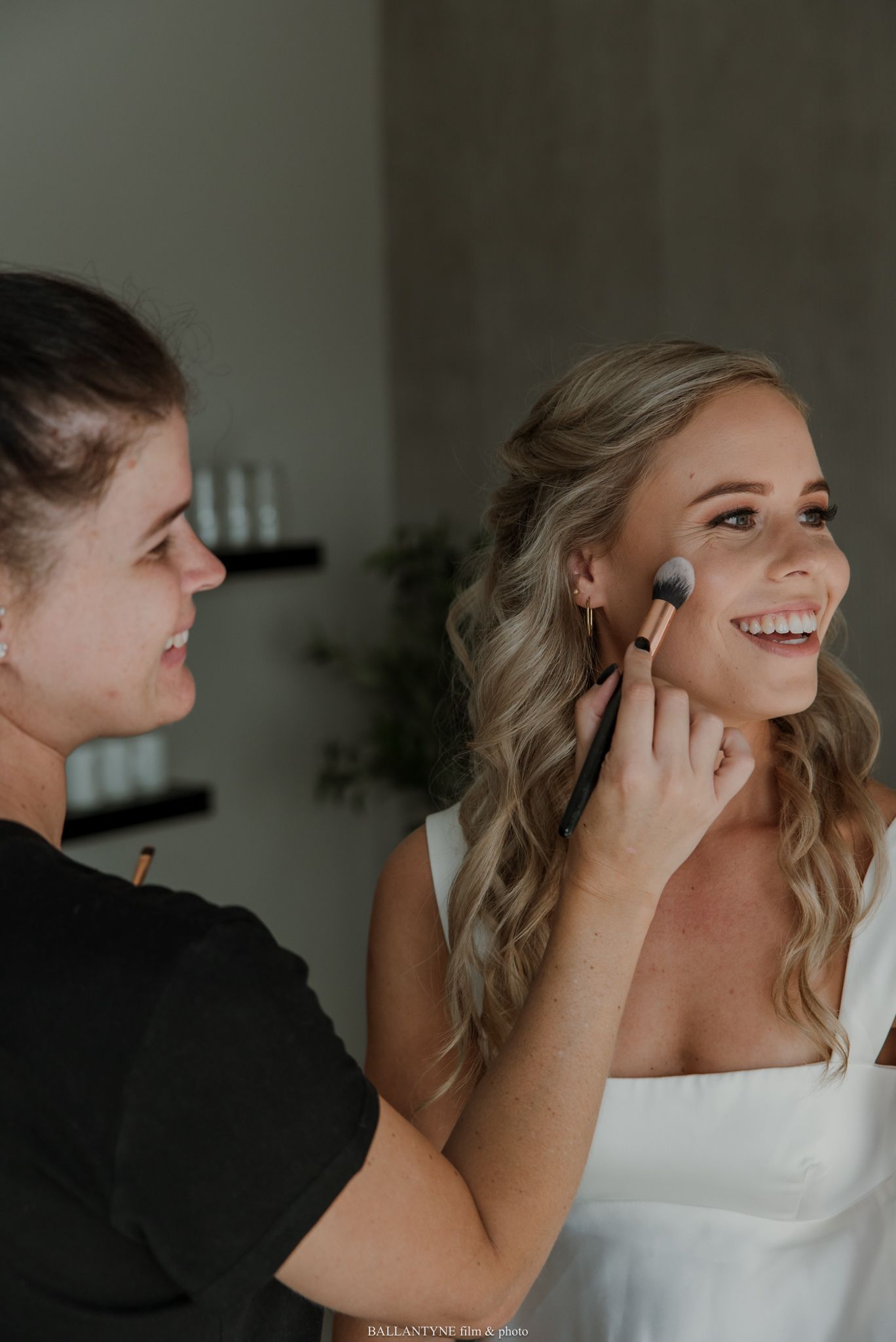 A makeup artist applies blush to a smiling bride in a white dress, preparing her for her wedding day.