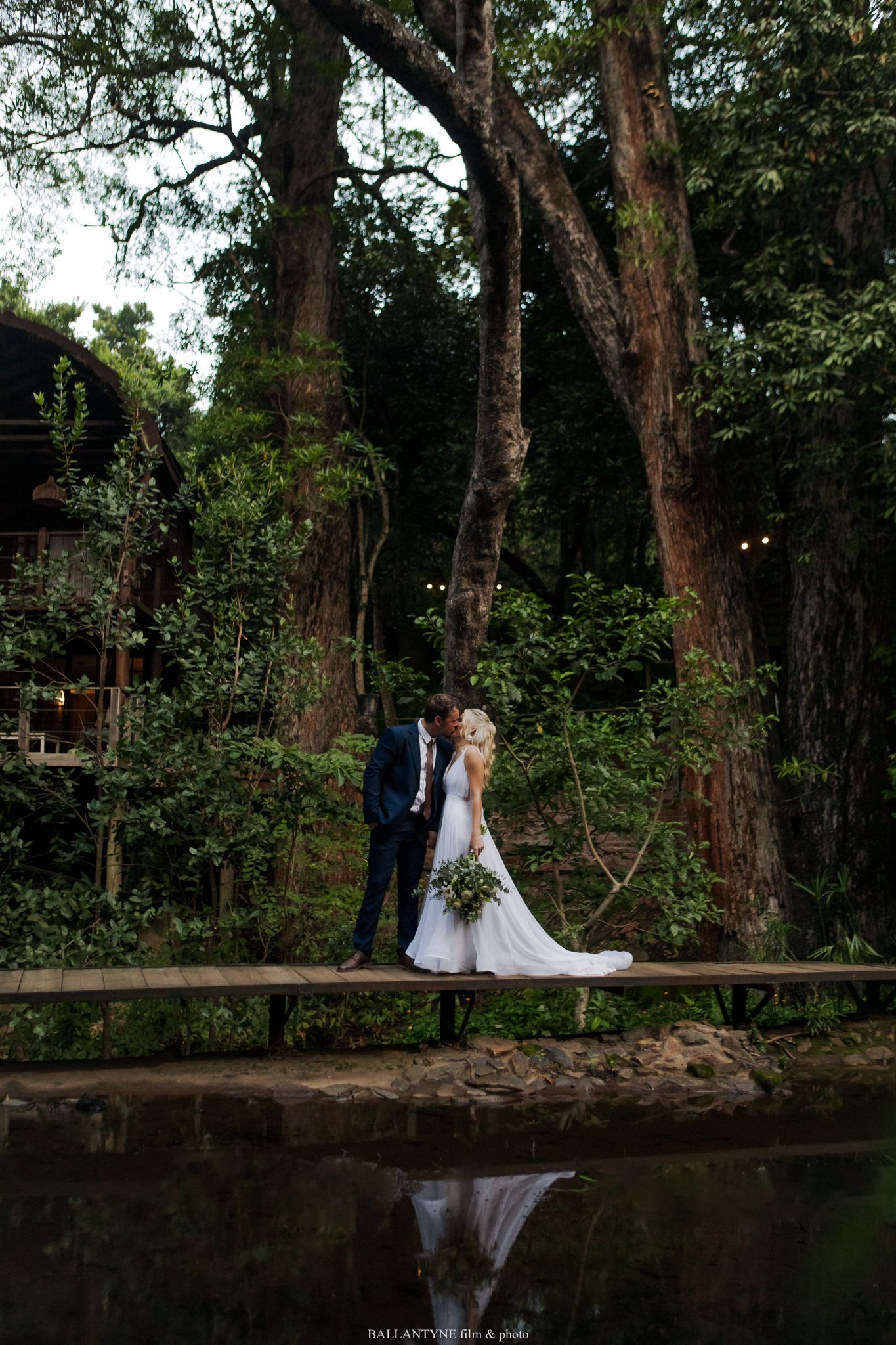 Wedding couple poses near reflective pond surrounded by tall trees in romantic forest setting.