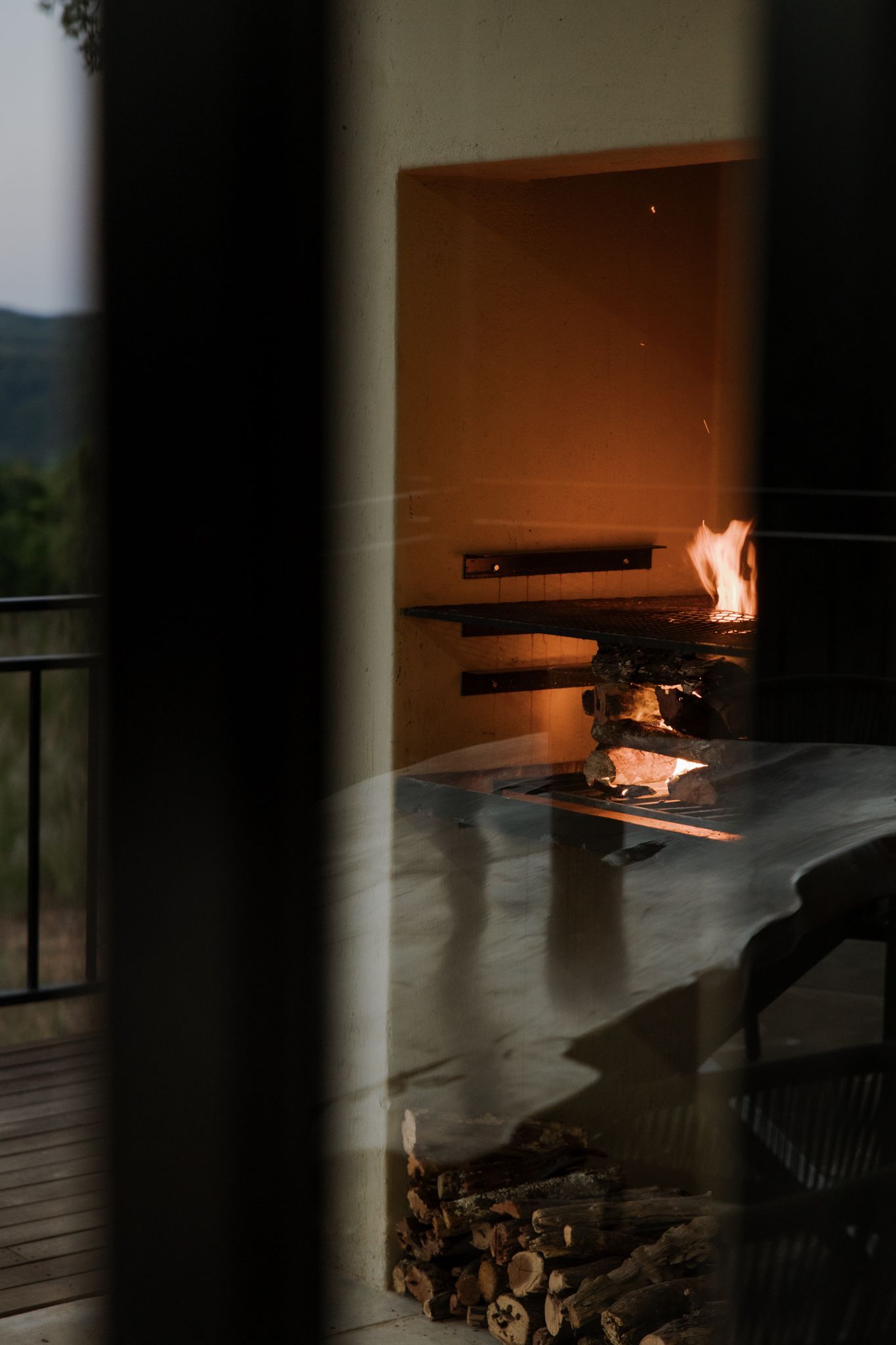A cozy fireplace with burning logs illuminates a balcony view at dusk through a glass door reflection.