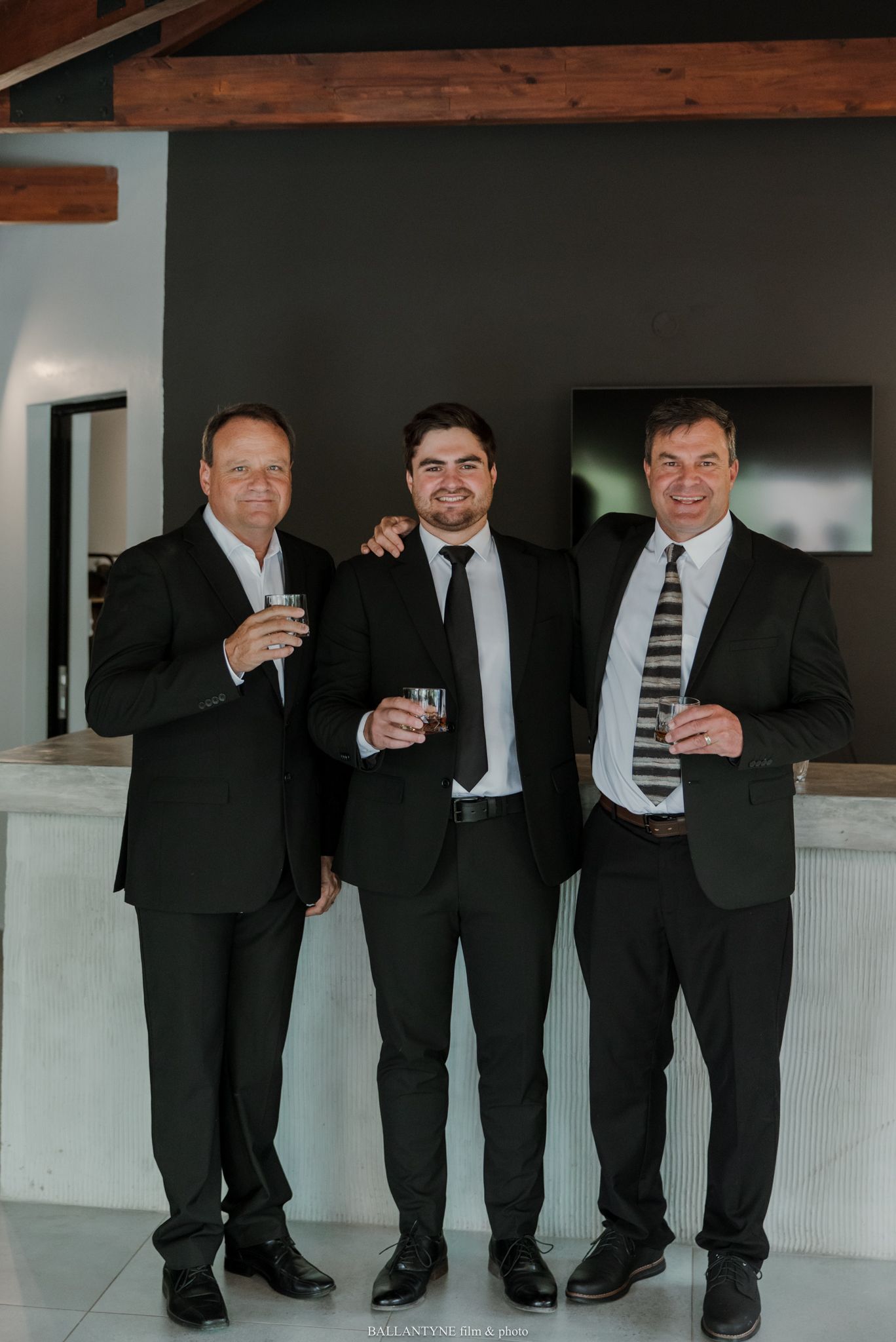Three men in black suits holding drinks pose for a photo at an elegant indoor venue.