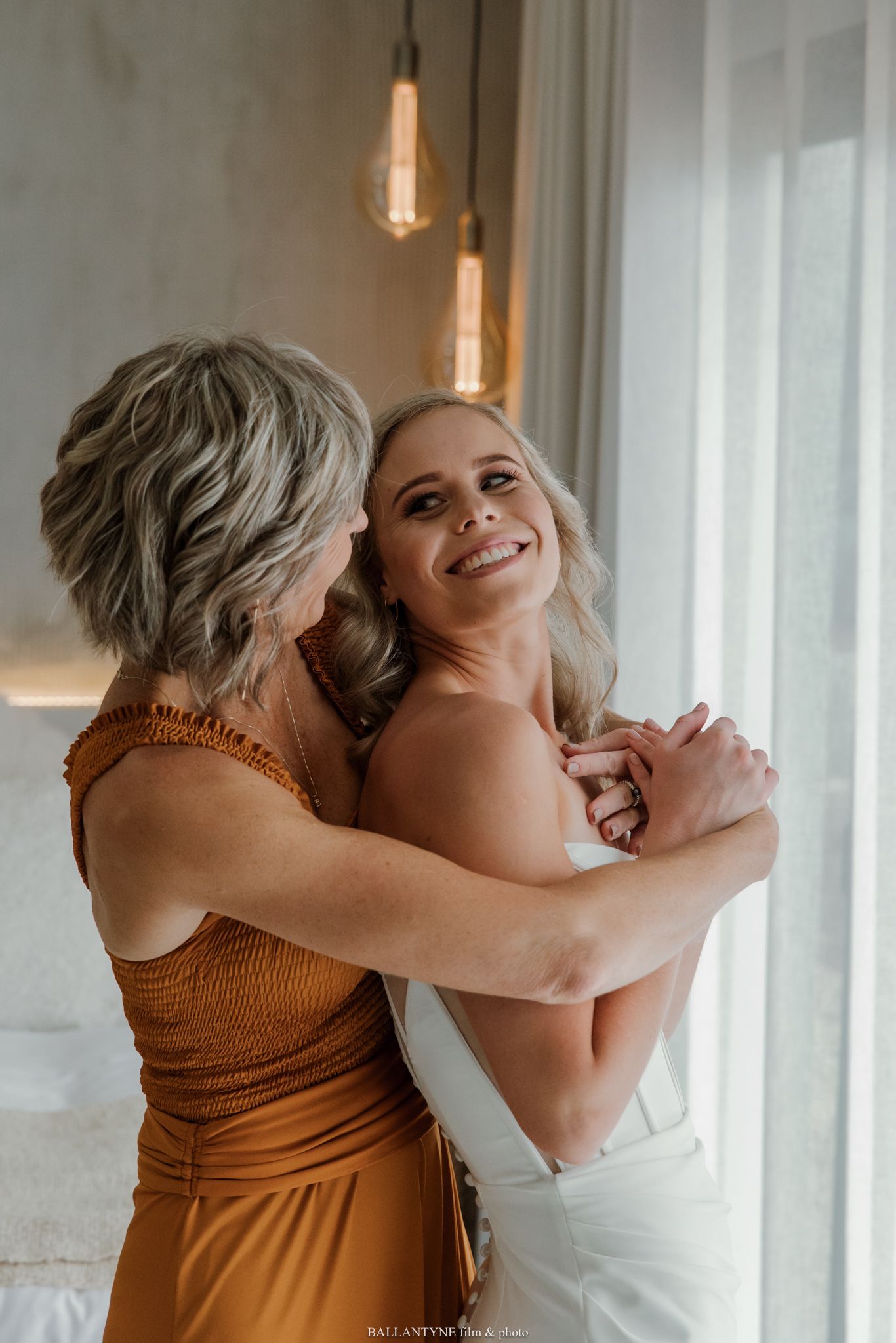 Two blonde women in elegant attire share a warm embrace near a sunlit window, one wearing a rust-colored dress.