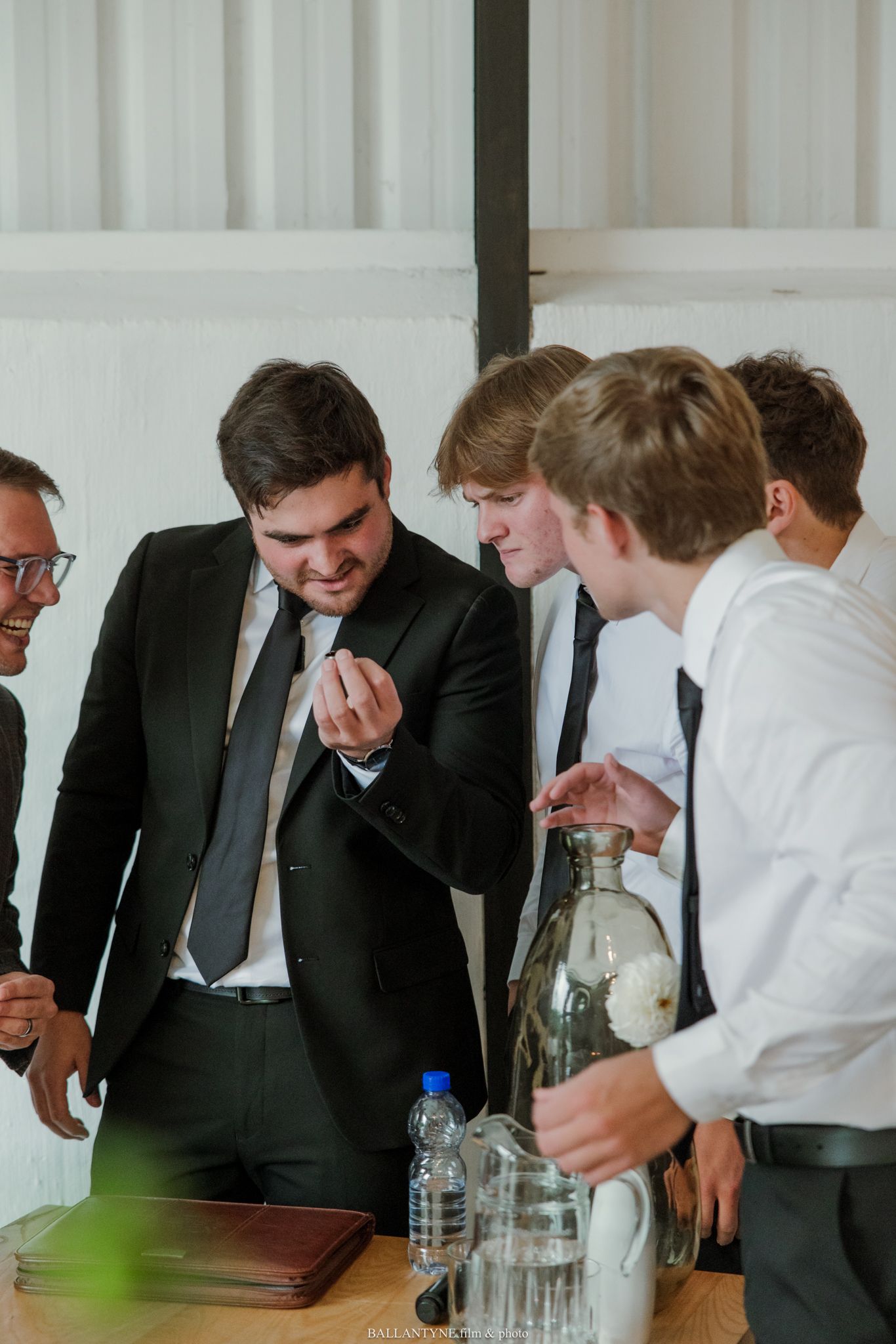 Group of young men in formal suits gathered around a table, examining something closely at an indoor event.