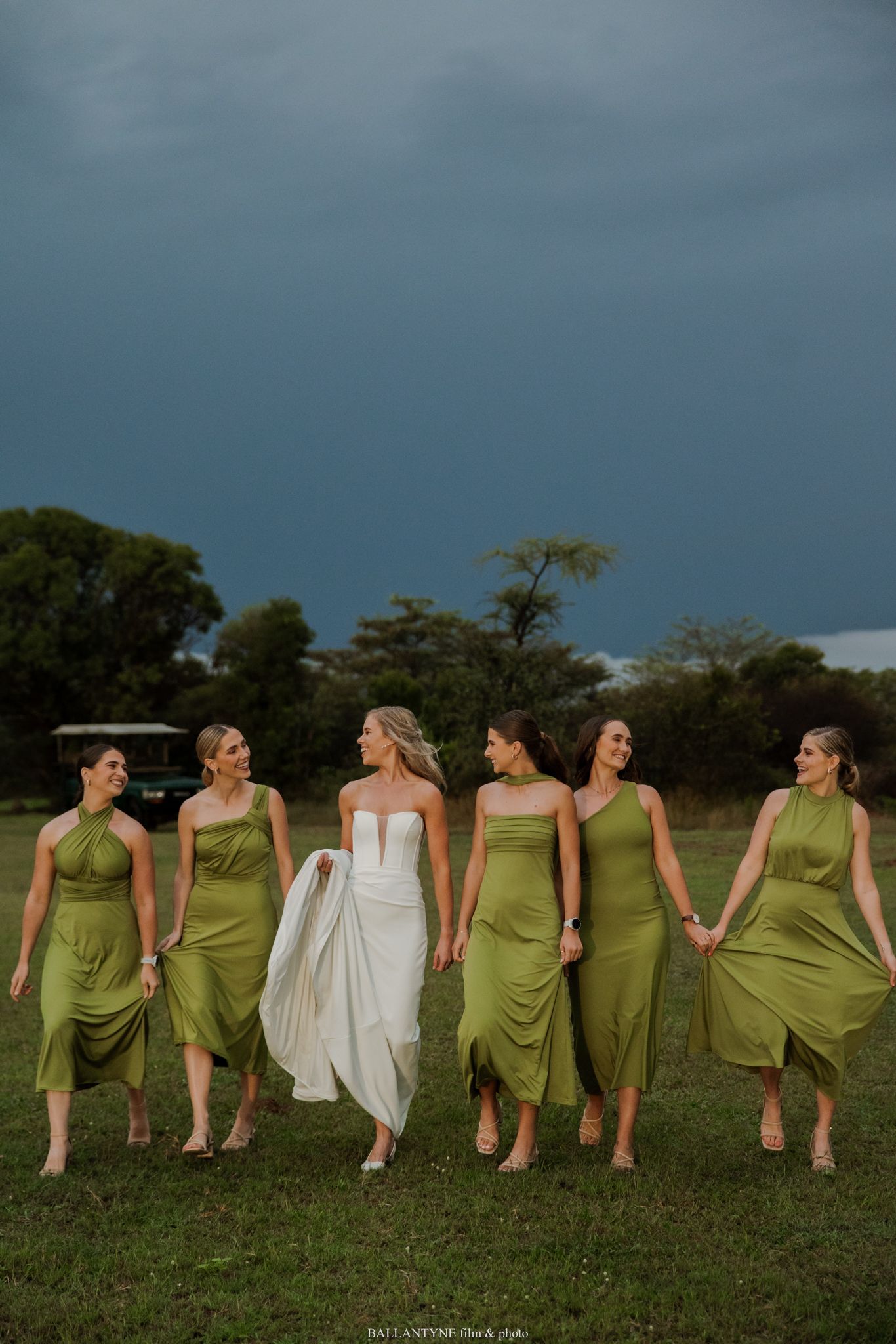 Bride in white gown walks with five bridesmaids in olive green dresses across a field under dramatic stormy skies.