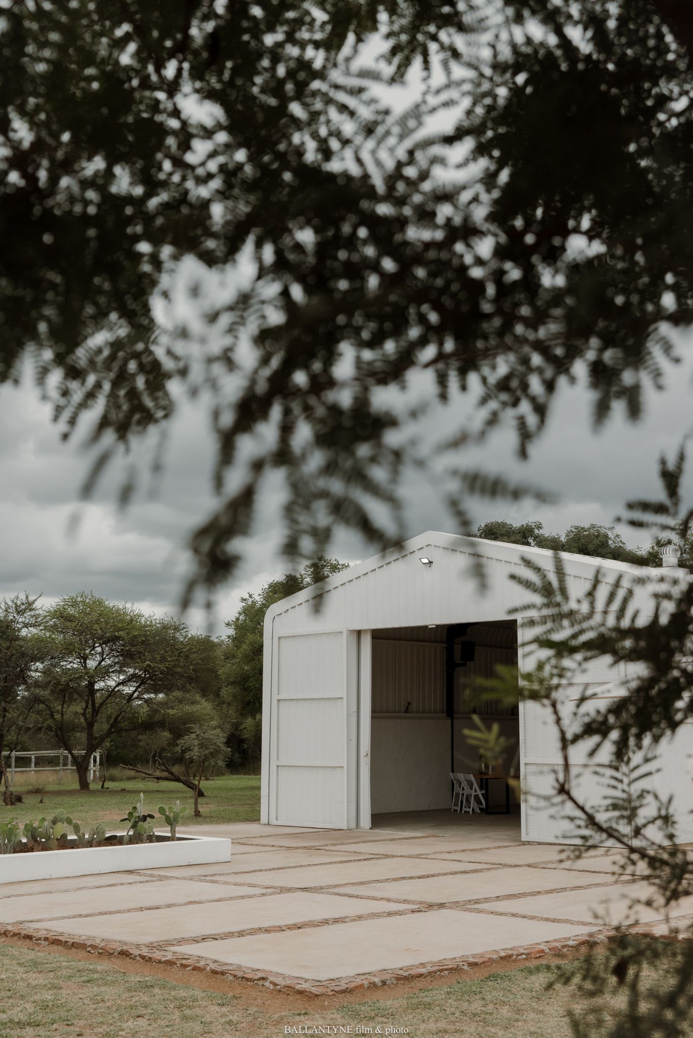 White barn venue with open doors on a paved outdoor area, framed by lush green trees under a cloudy sky.