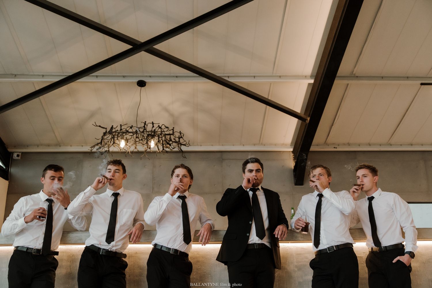 Six groomsmen in white shirts and black ties toasting drinks at a bar during a wedding celebration.