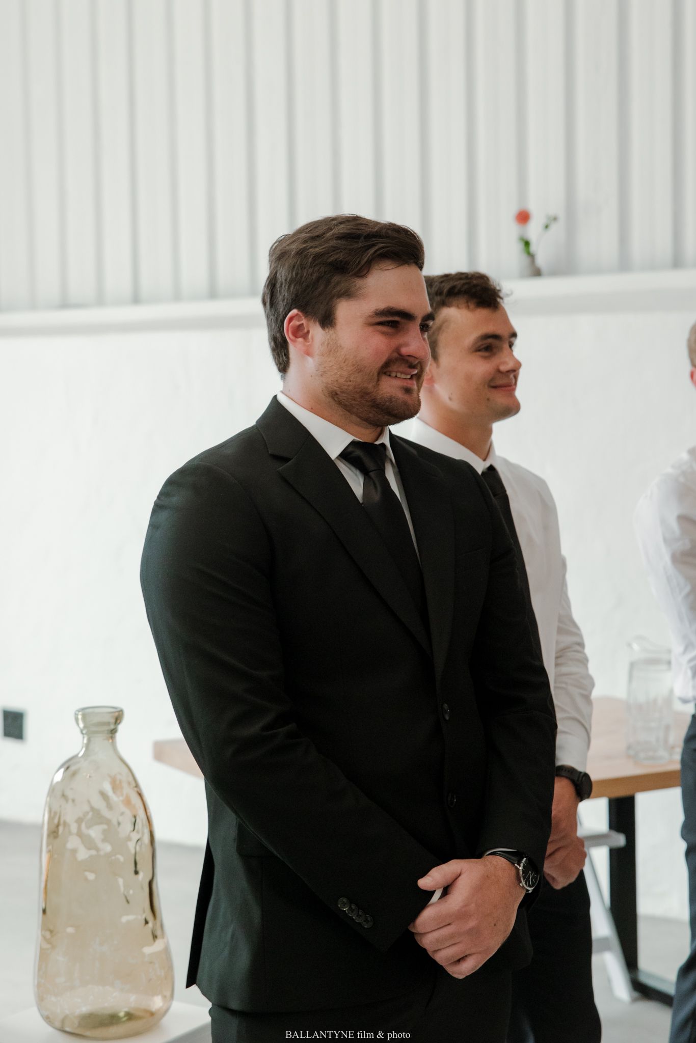 Two men in black suits smiling at a wedding ceremony in a bright white venue.
