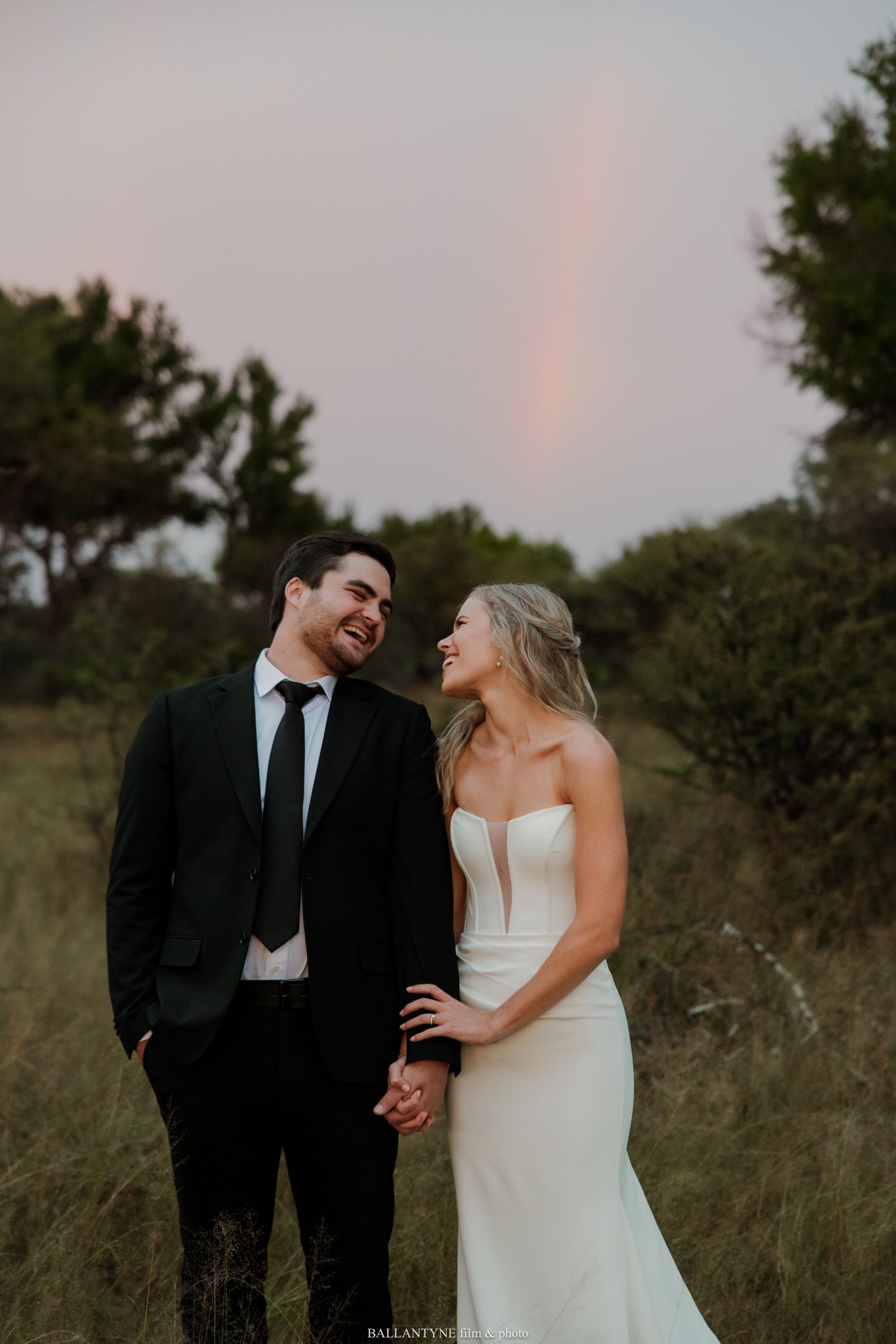 Laughing bride and groom hold hands in a field with a rainbow arching in the sky behind them.