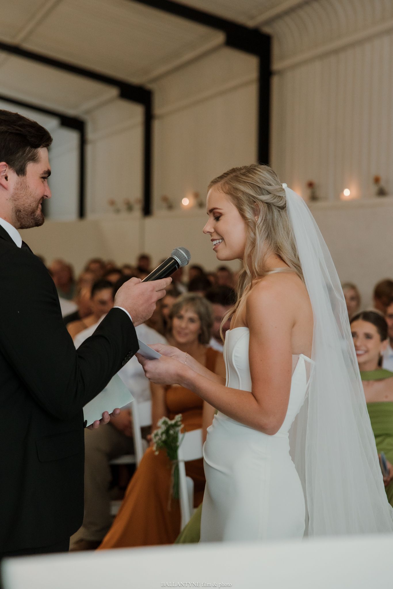Bride smiling while exchanging vows with groom at elegant indoor wedding ceremony with guests seated in background.