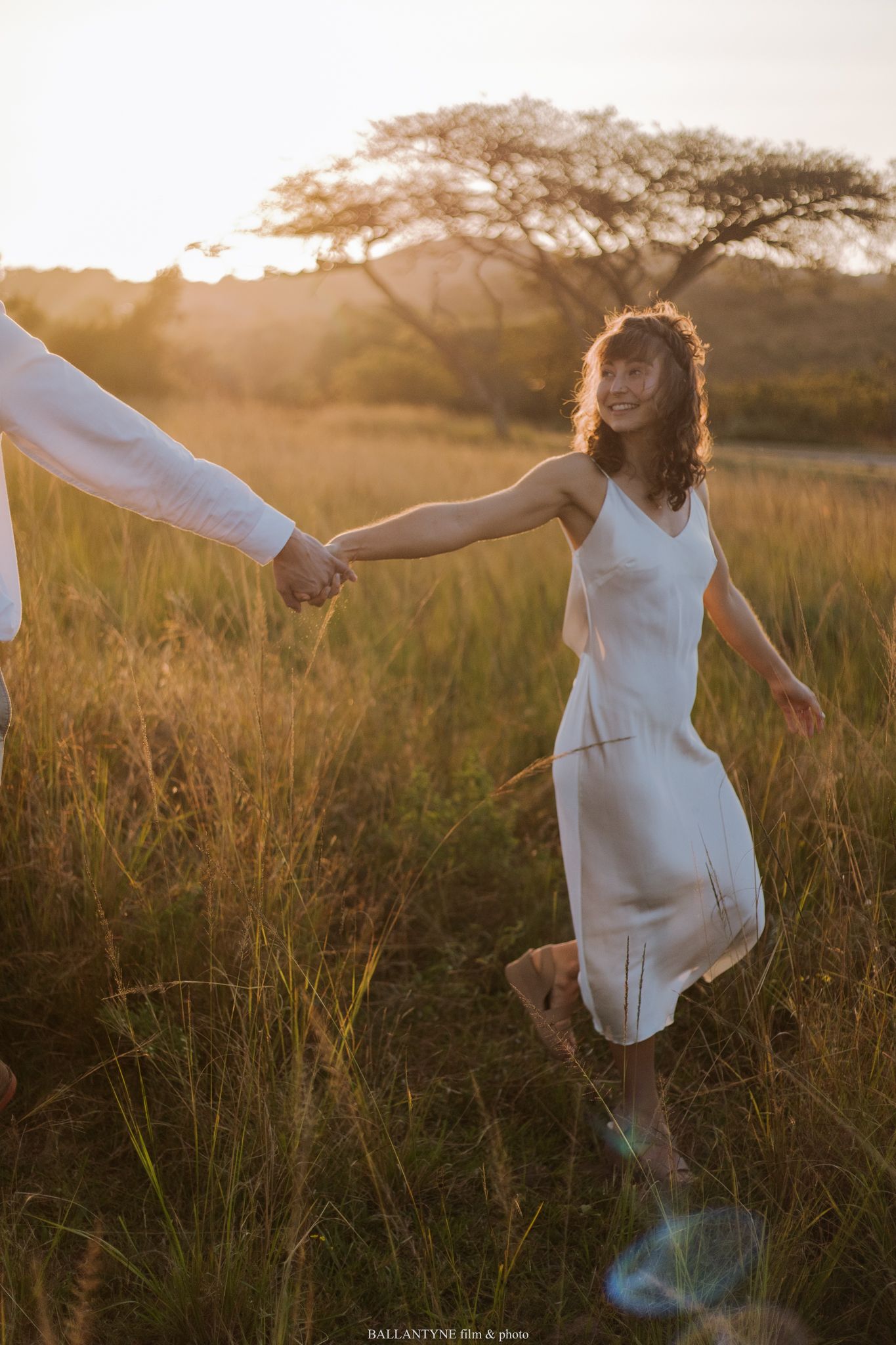 A person in a white dress twirls gracefully through a sunlit field of tall grass during golden hour.