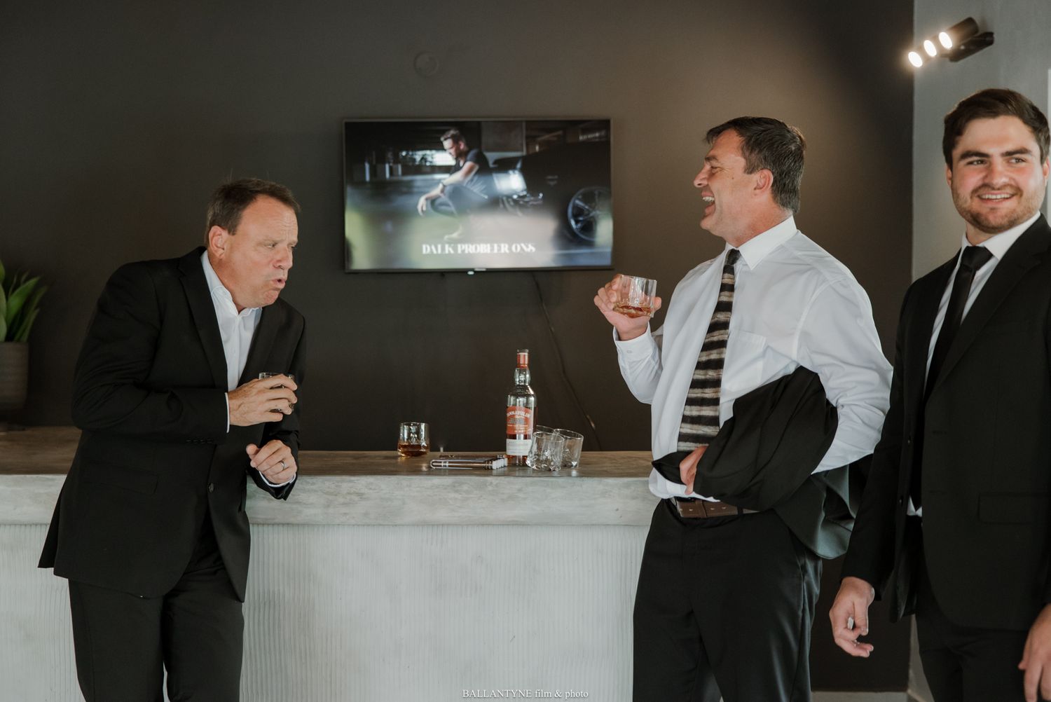 Three men in suits socializing at a modern bar counter with drinks and a TV screen in the background.