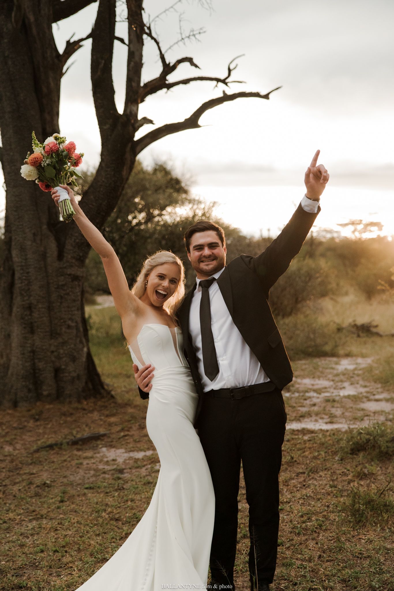 Joyful newlyweds celebrate outdoors at sunset, bride in white gown holding bouquet, groom in black suit, arms raised.
