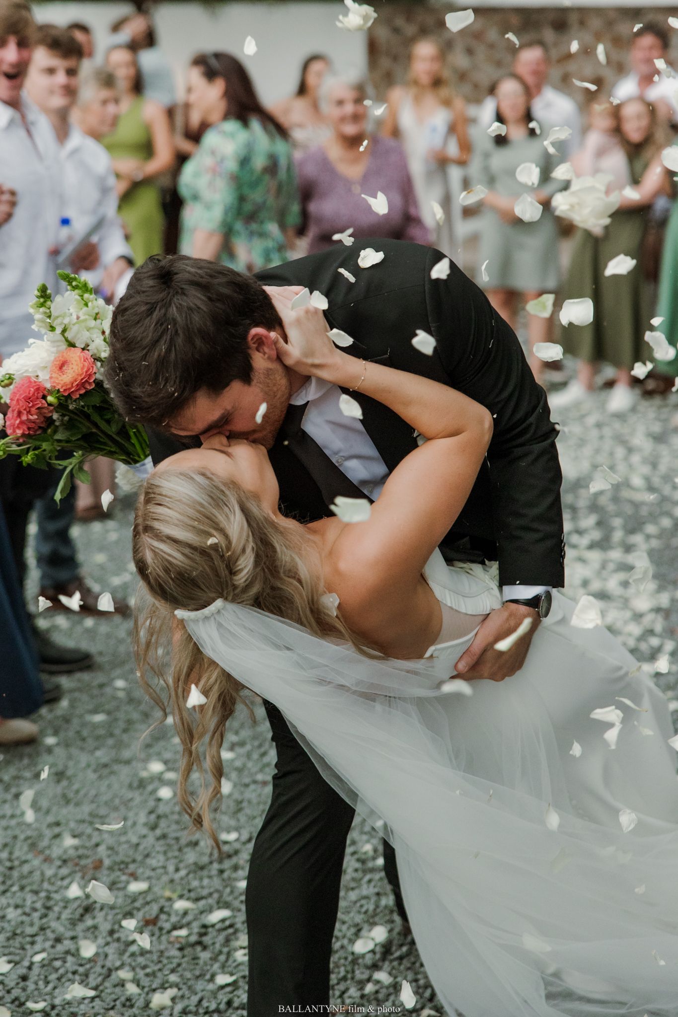 Bride and groom share a romantic dip kiss surrounded by guests tossing white flower petals at outdoor wedding celebration.