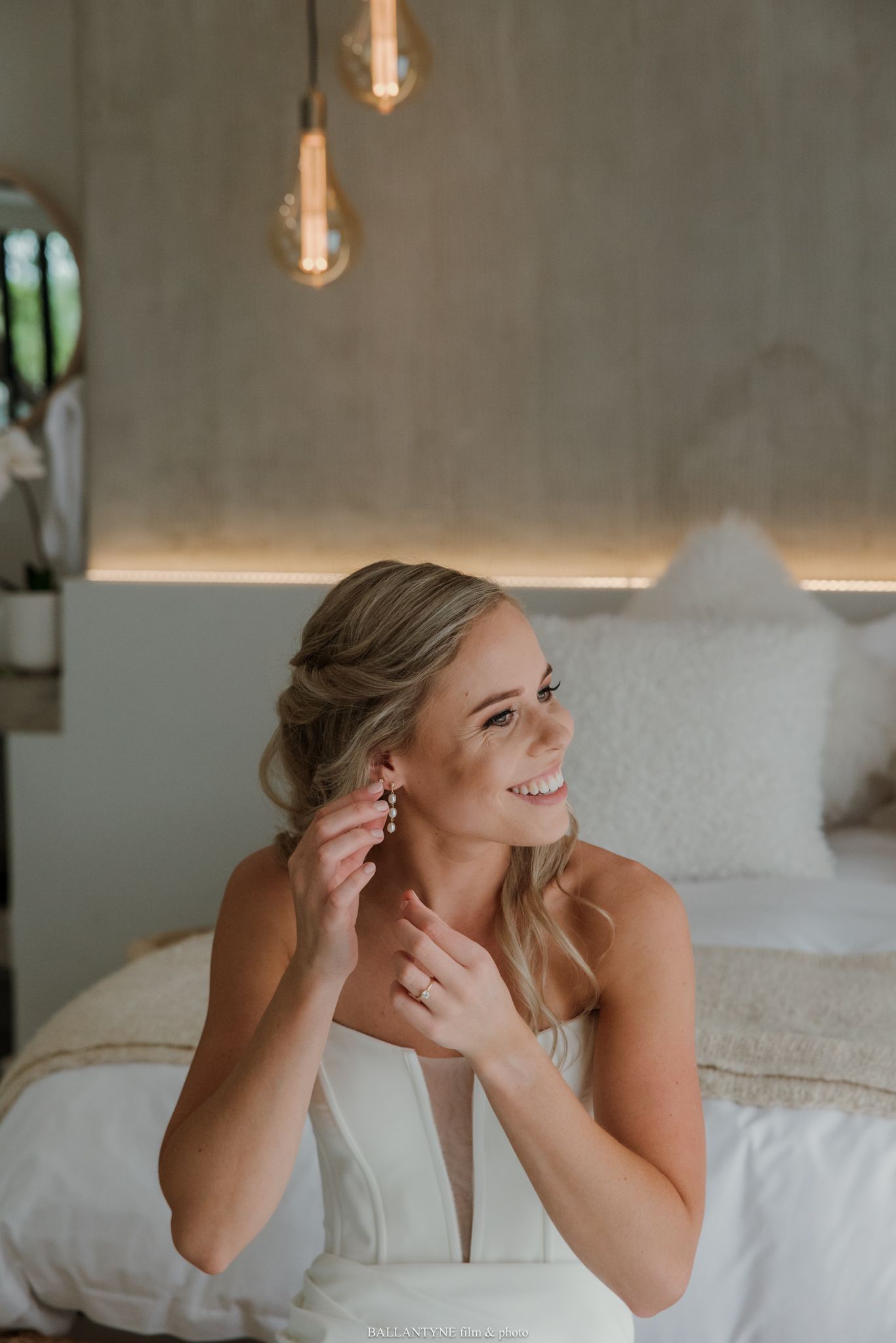 Smiling bride in white dress putting on earrings while sitting on bed with warm Edison bulb lighting.
