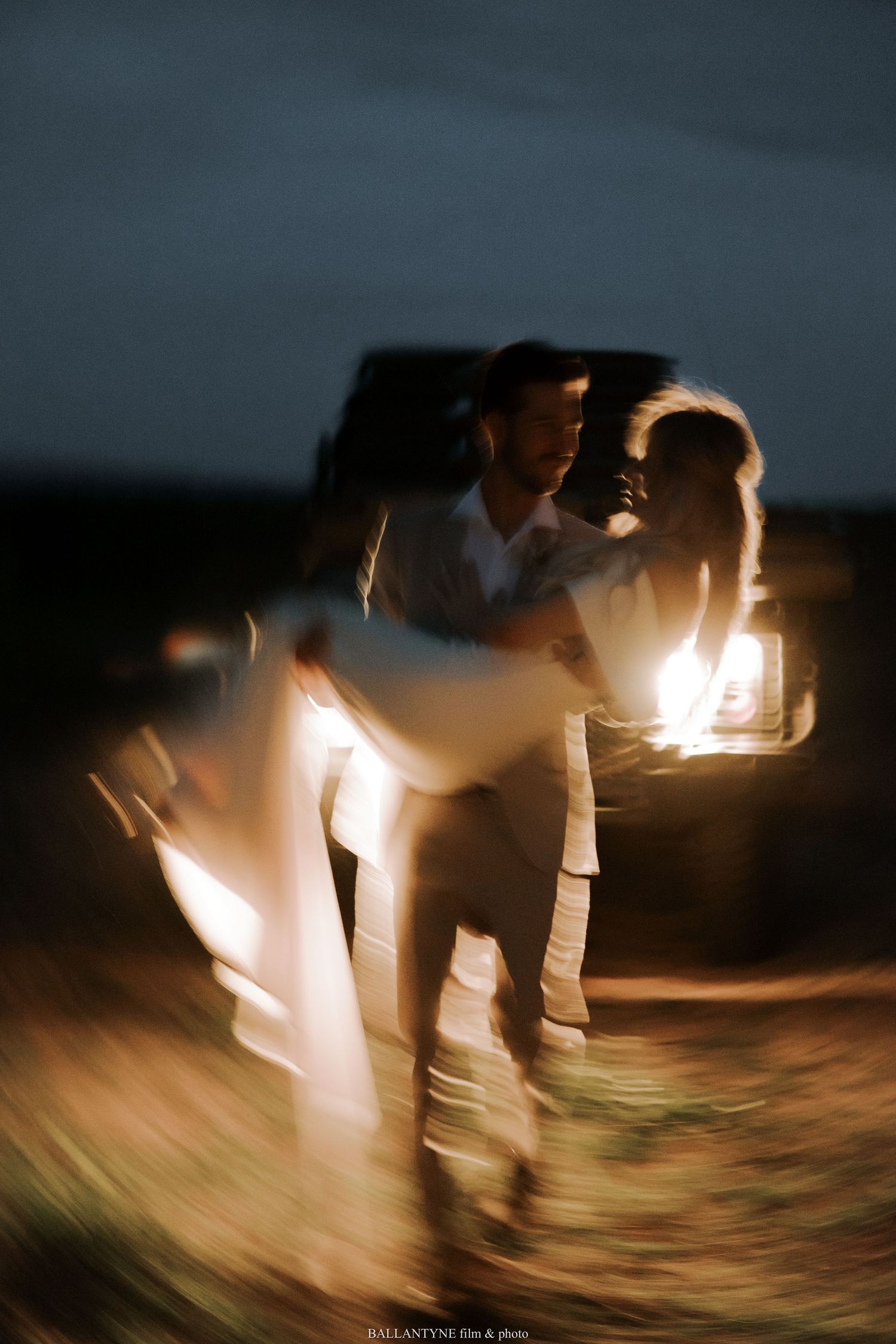 Artistic motion blur wedding photo with couple dancing in truck headlights at night.
