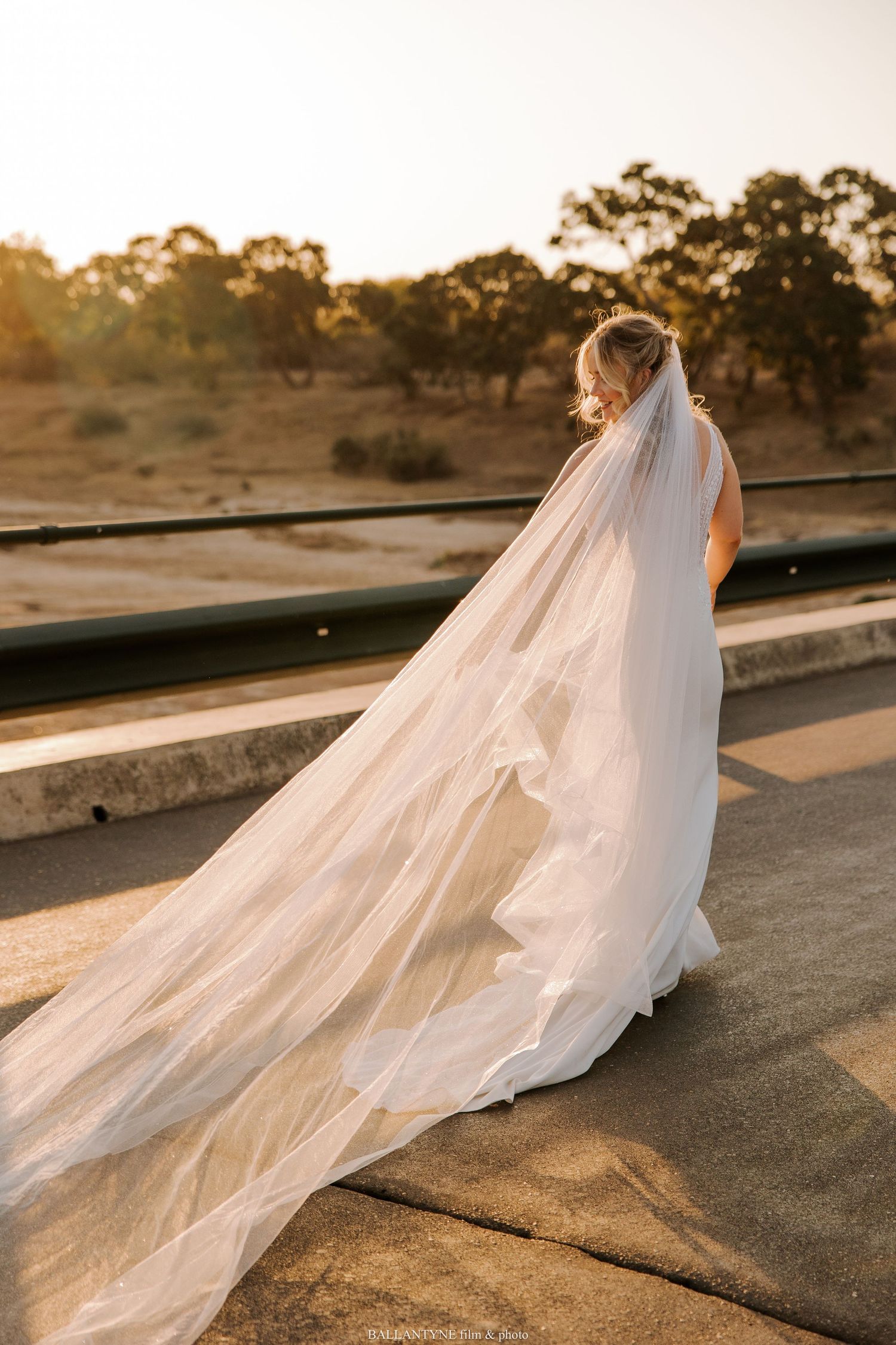 Long cathedral veil trails behind wedding dress against sunset landscape.