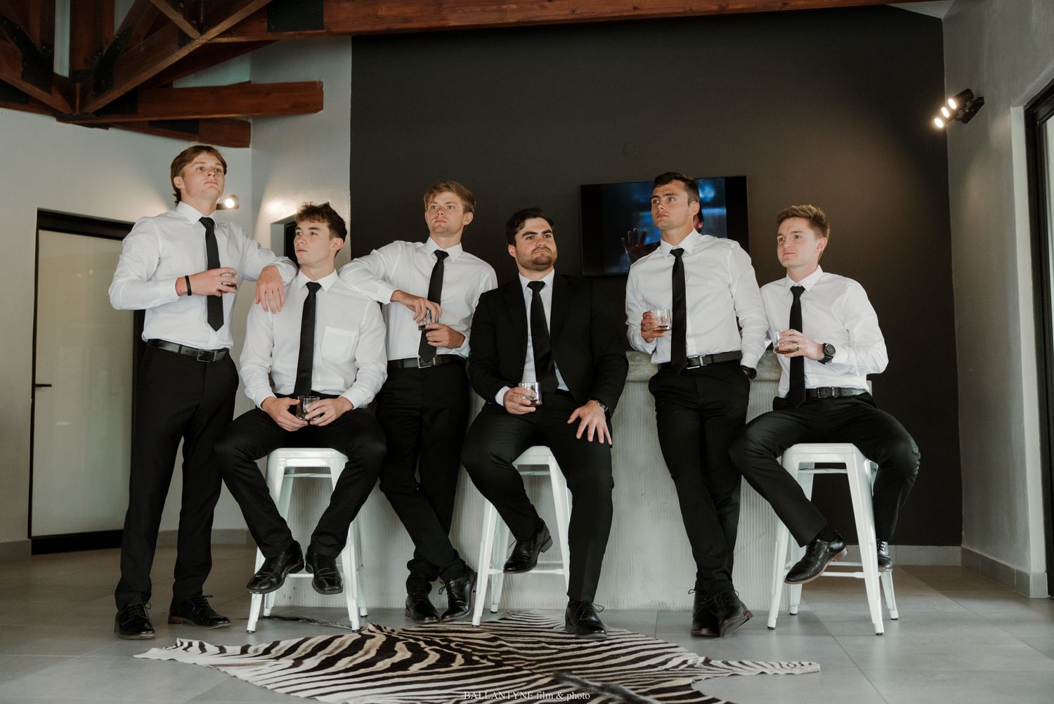 Six young men in white shirts and black ties pose confidently in a modern studio space with a zebra-print rug.