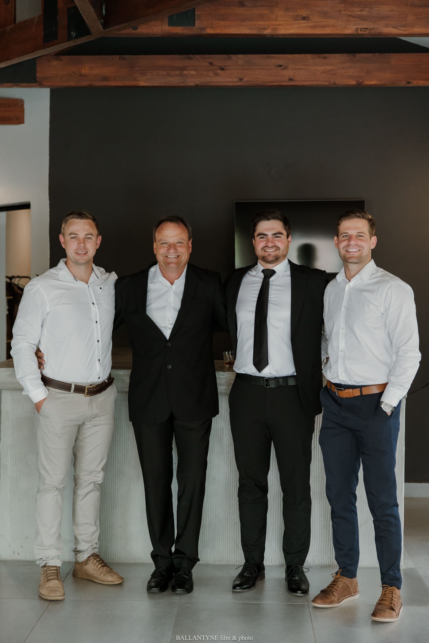 Four men in formal attire smiling together indoors, posing for a group photo at what appears to be a special event.