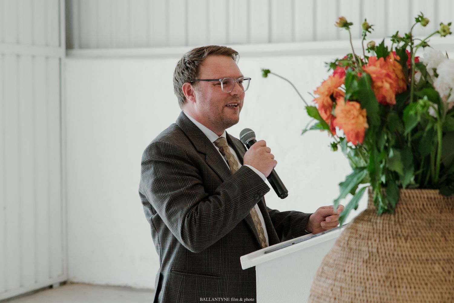 Man in suit speaking into microphone at podium with orange floral arrangement at formal event.