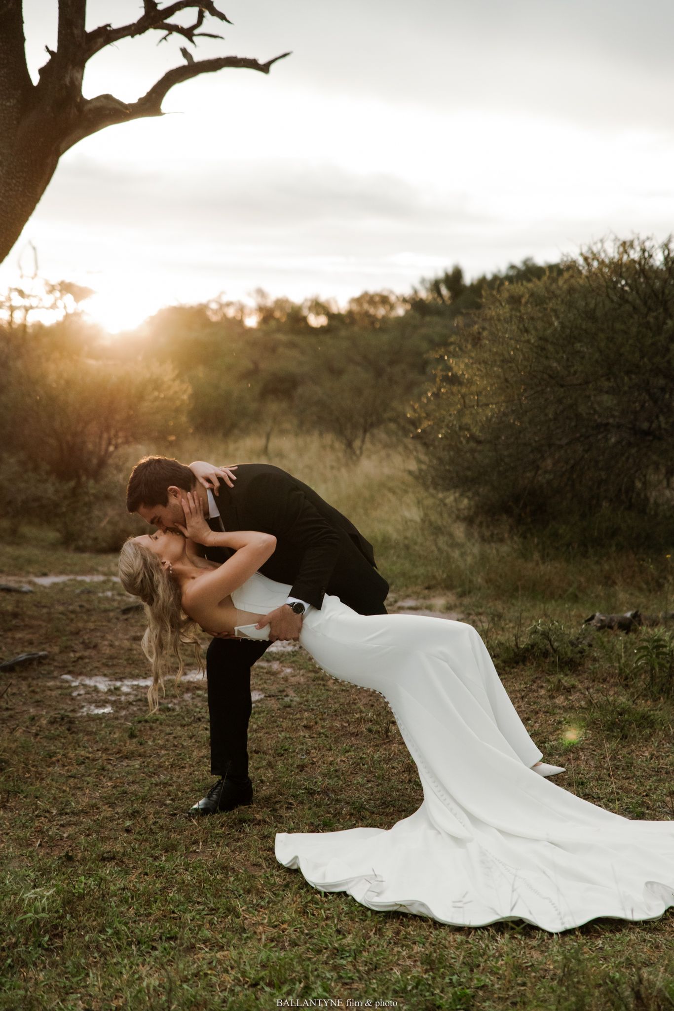 Bride in white gown dips with groom in black suit at golden sunset in lush outdoor wedding portrait.