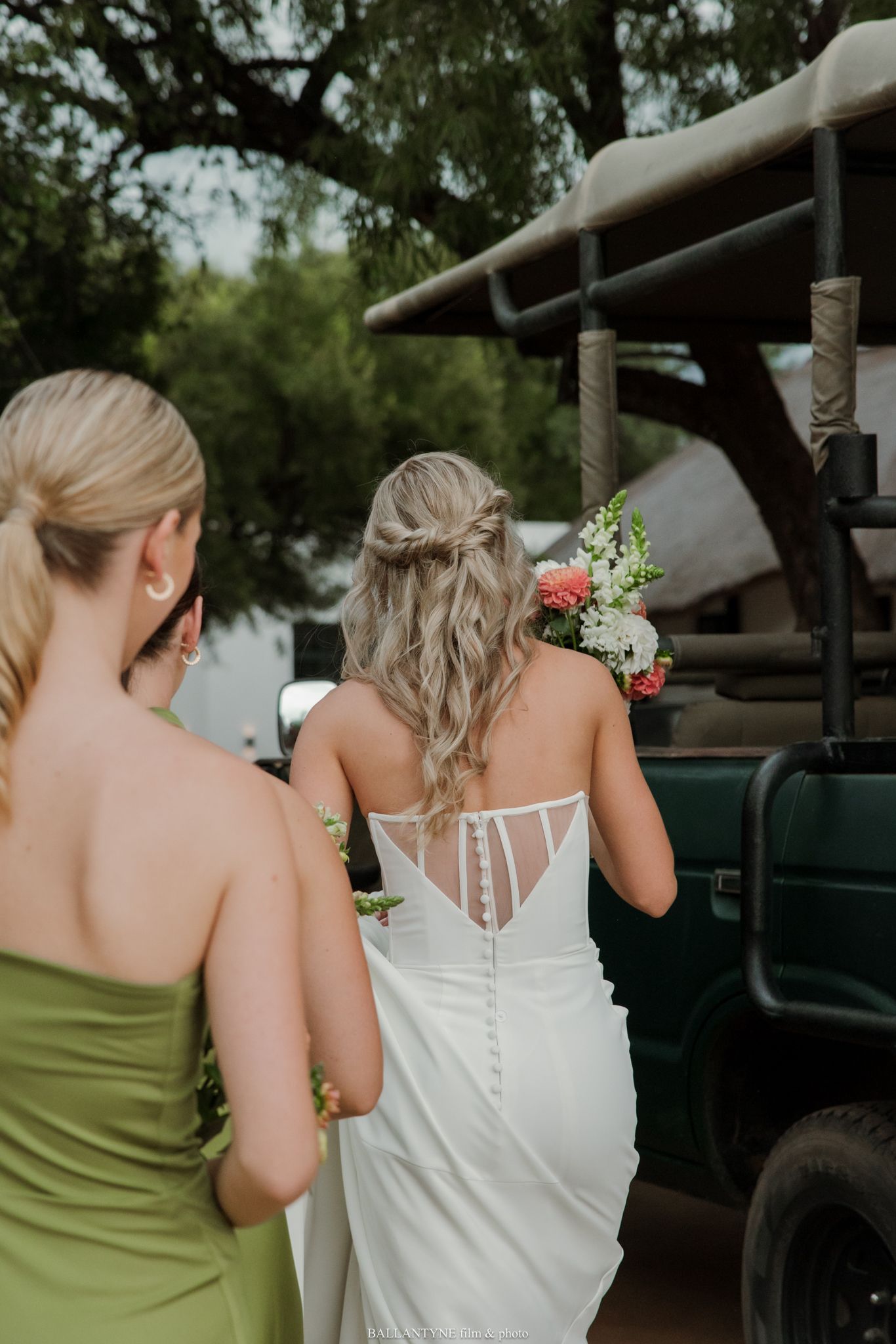 Bride in white gown with floral bouquet walks with bridesmaid in green dress toward safari vehicle outdoors.