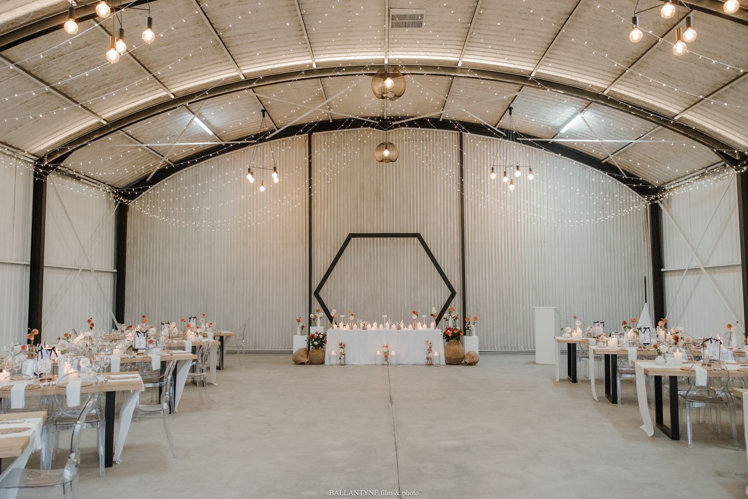 Elegant wedding reception in an industrial barn venue with hexagon arch, string lights, and white-draped tables.