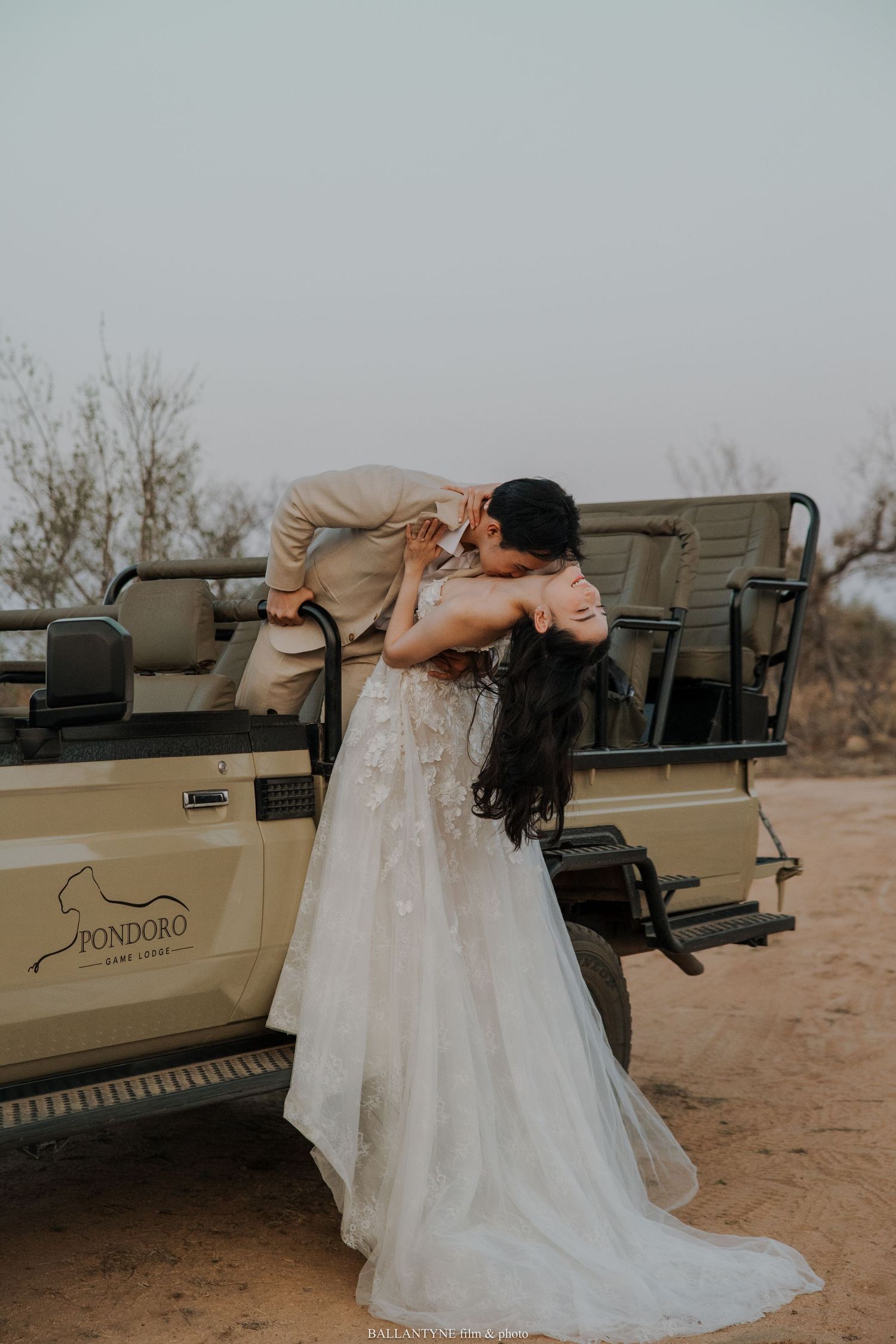A romantic wedding couple embraces in front of a safari vehicle on a dusty road during a destination wedding.