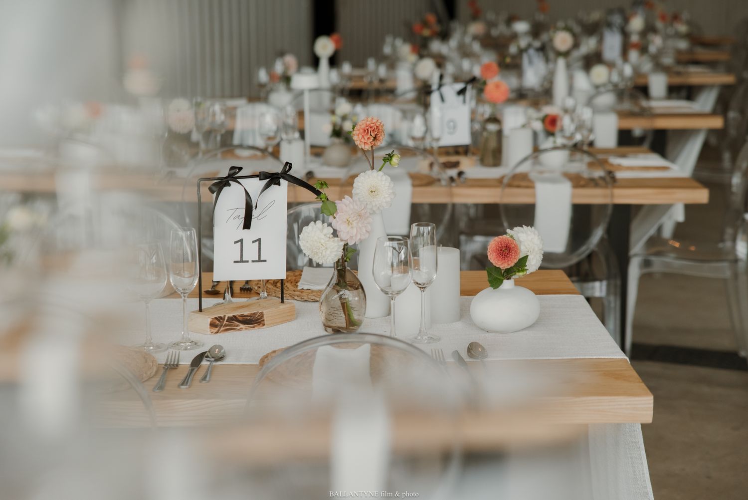 Elegant wedding reception tables with white floral centerpieces, table number 11, and rustic wooden tables in a bright venue.