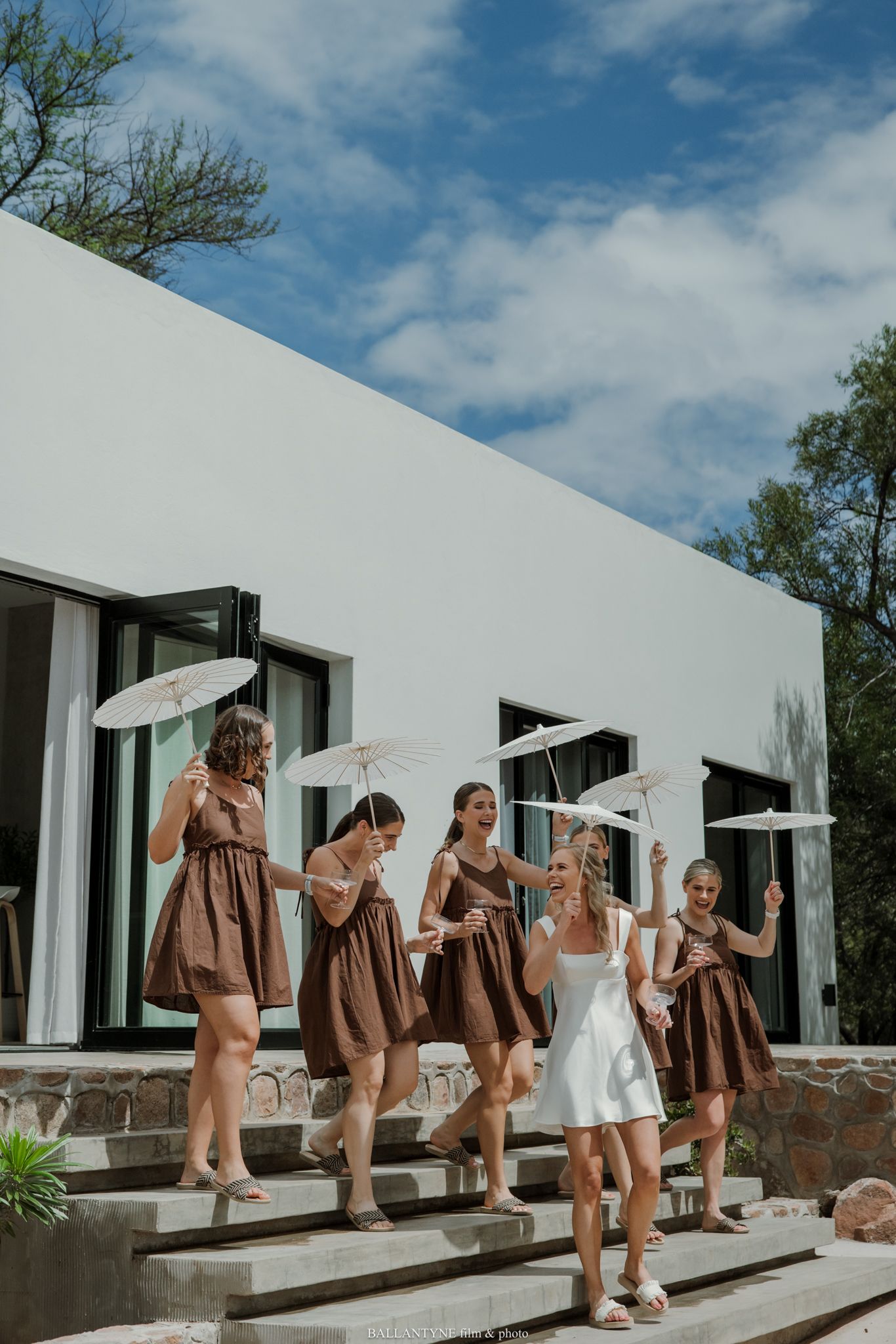 Bride in white dress with bridesmaids in brown dresses holding parasols on steps outside a modern white building.
