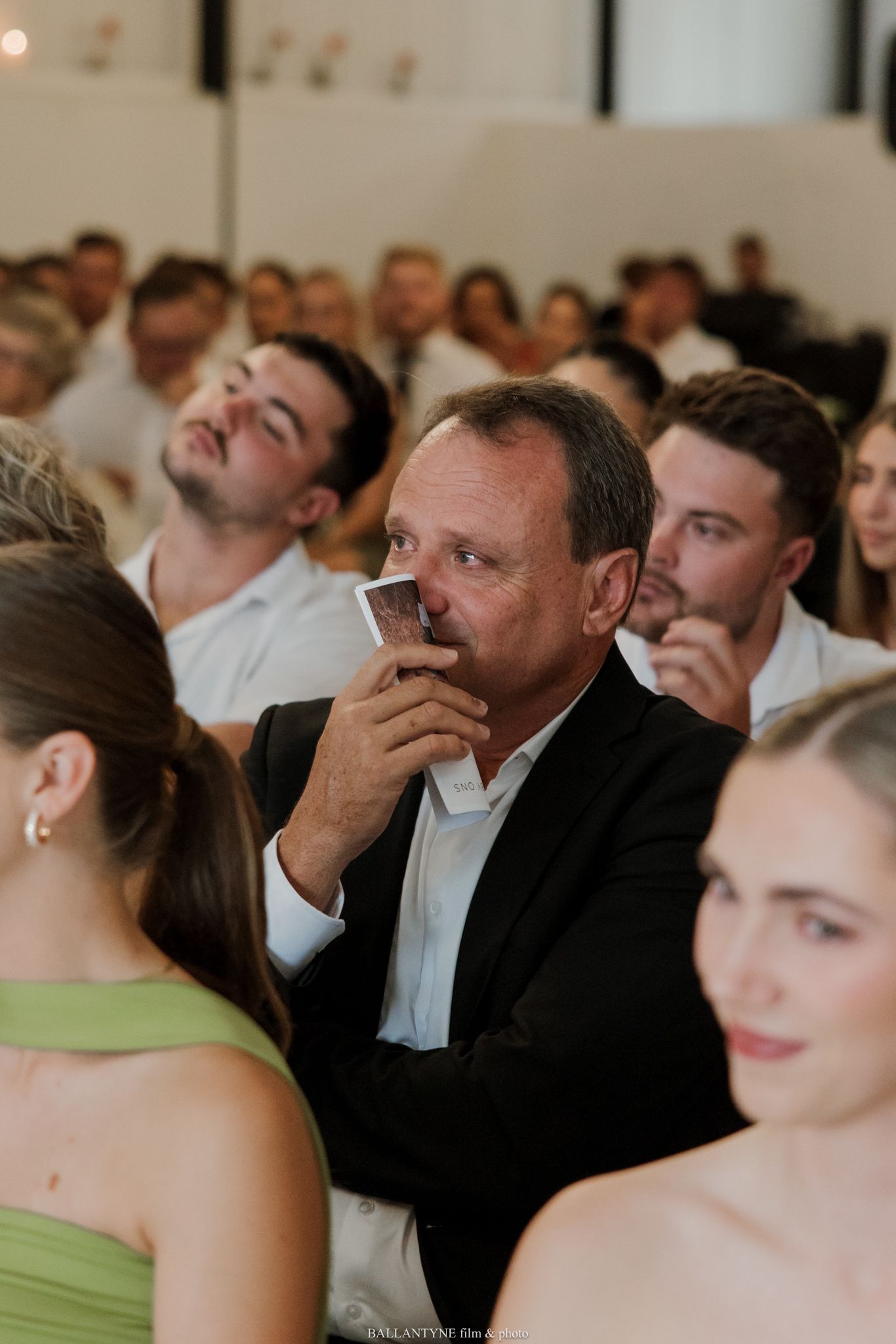 A man in a black suit holds a card to his face while seated among wedding guests in a bright venue.