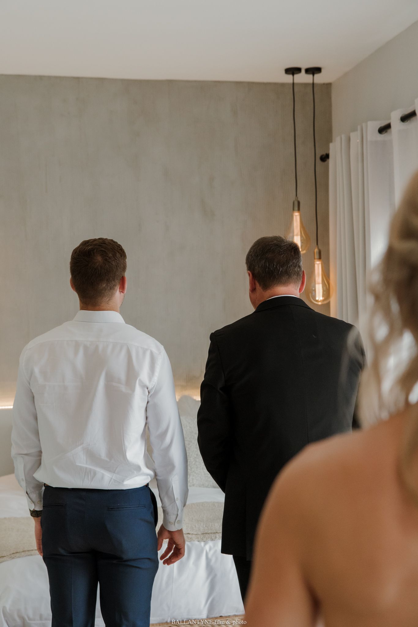 Two men in formal attire stand with backs turned in an elegant room with pendant lights, as a woman watches nearby.