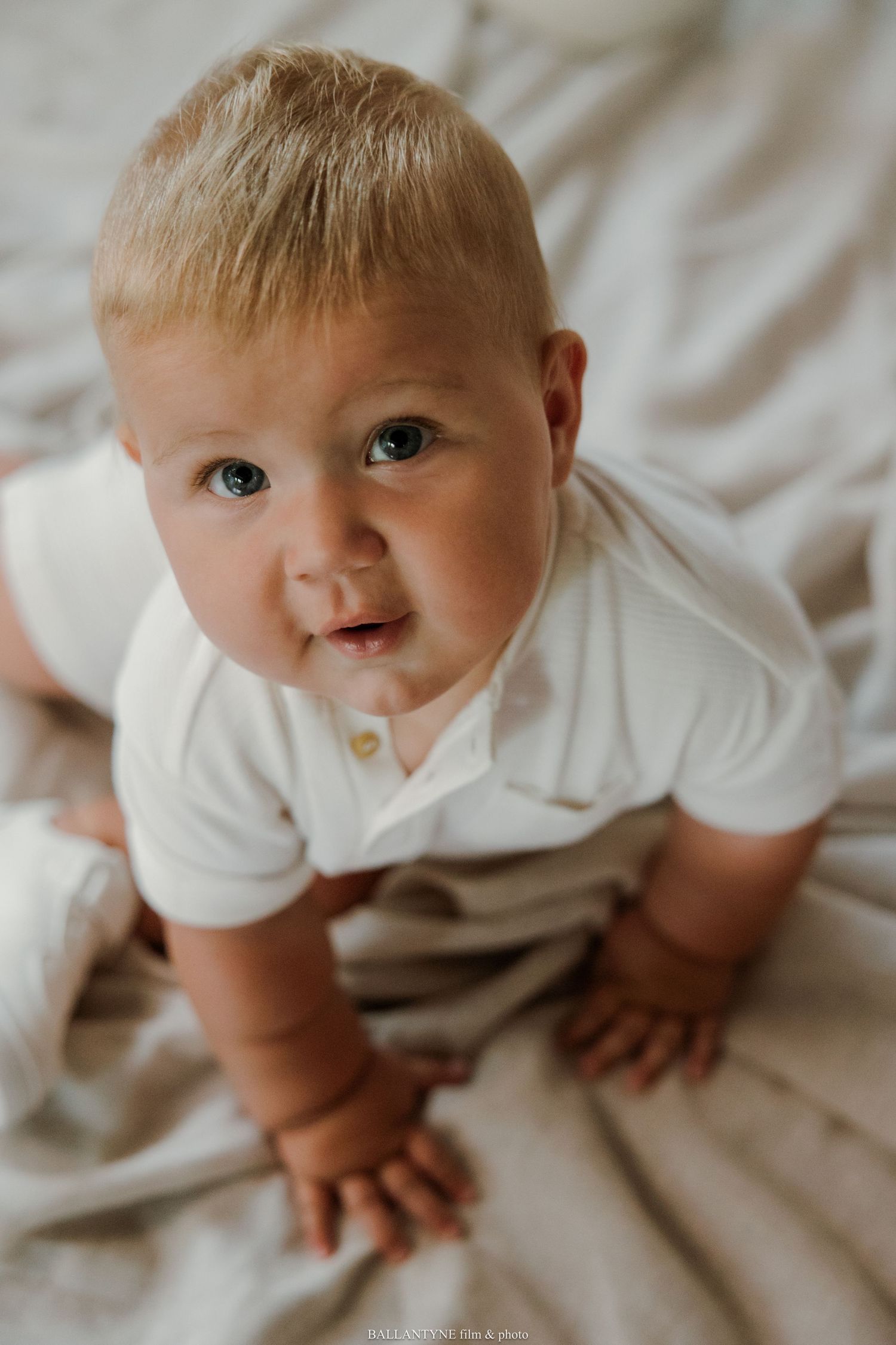 Baby in white outfit crawling on white bedding.