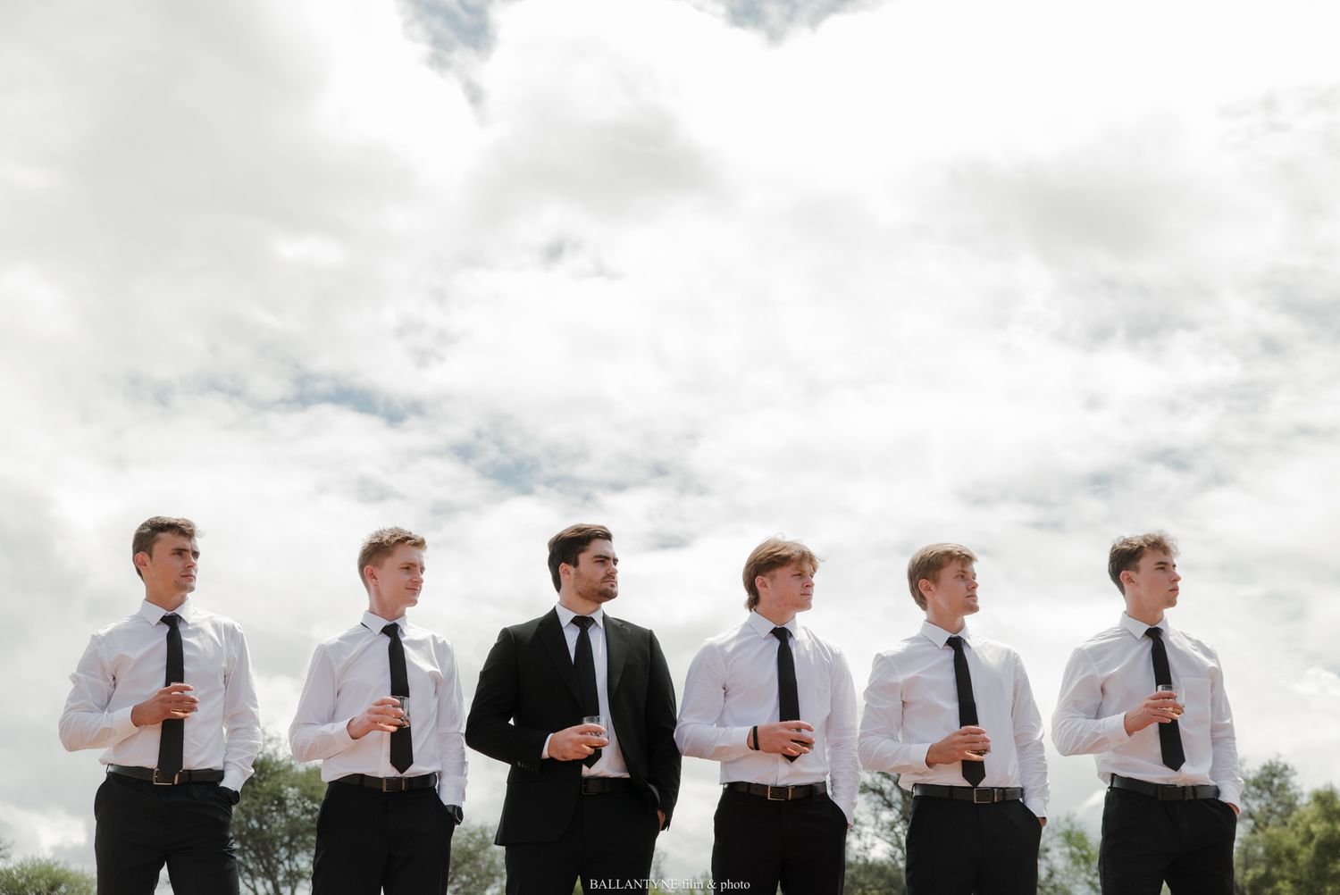 Groom in black suit stands with six groomsmen in white shirts and black ties under cloudy sky.