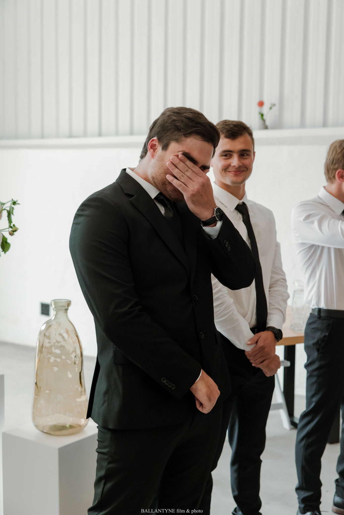 Emotional groom covering his face during wedding ceremony, standing beside smiling groomsmen in black suits.