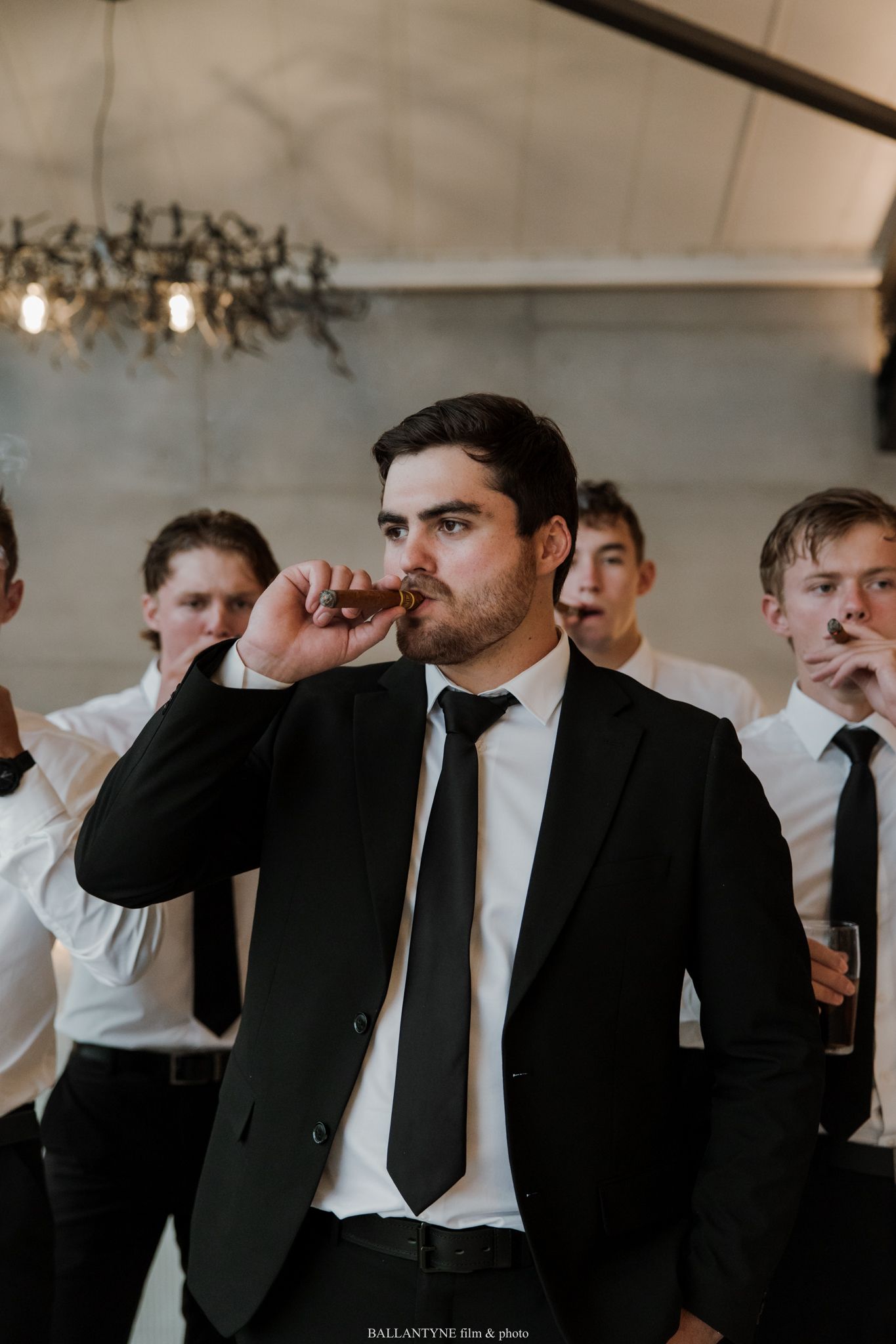 A groom in a black suit and tie smokes a cigar with groomsmen in white shirts during a wedding celebration.