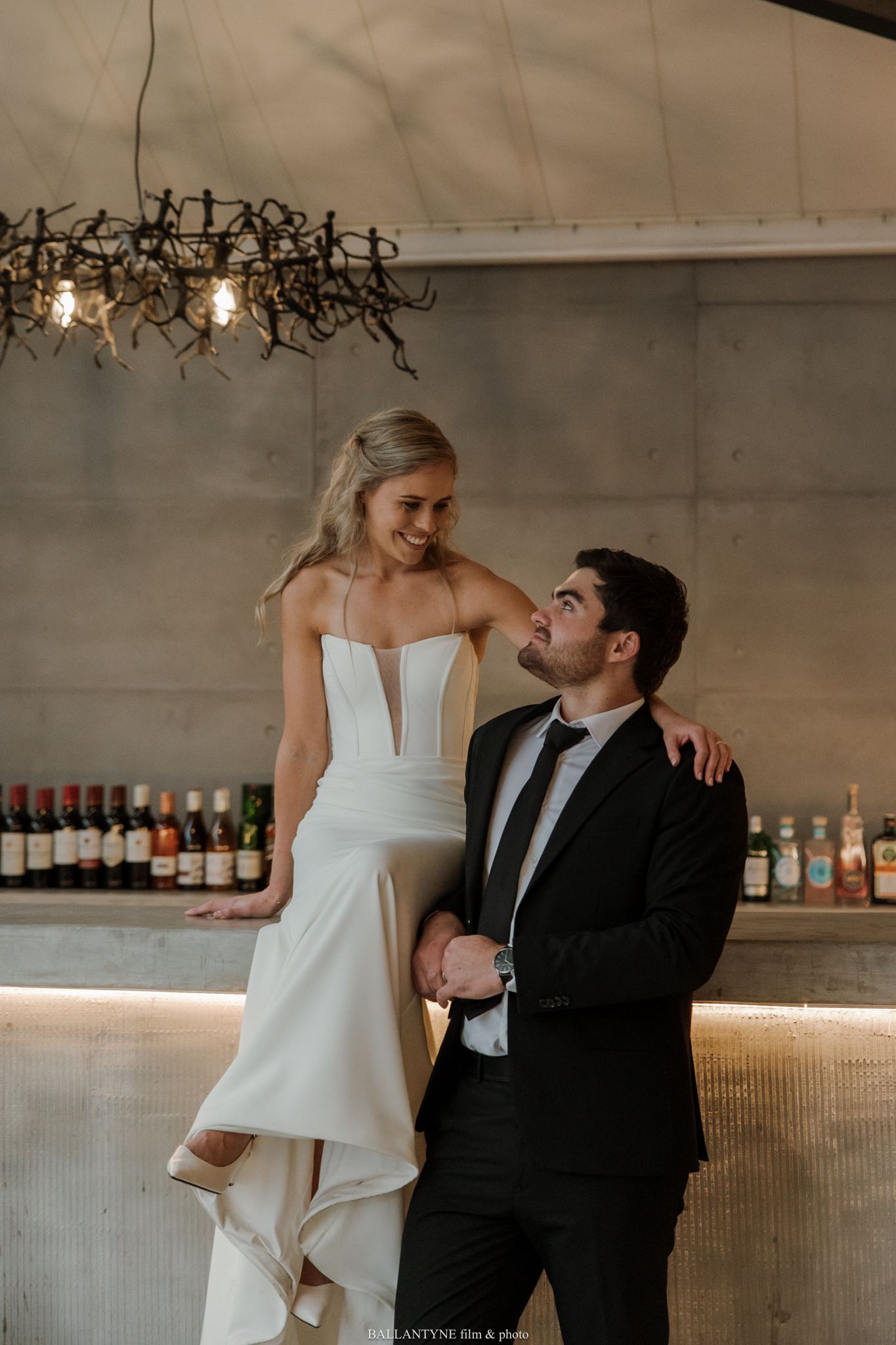 A bride in a white gown and groom in a black tuxedo share a romantic moment at an elegant bar venue.