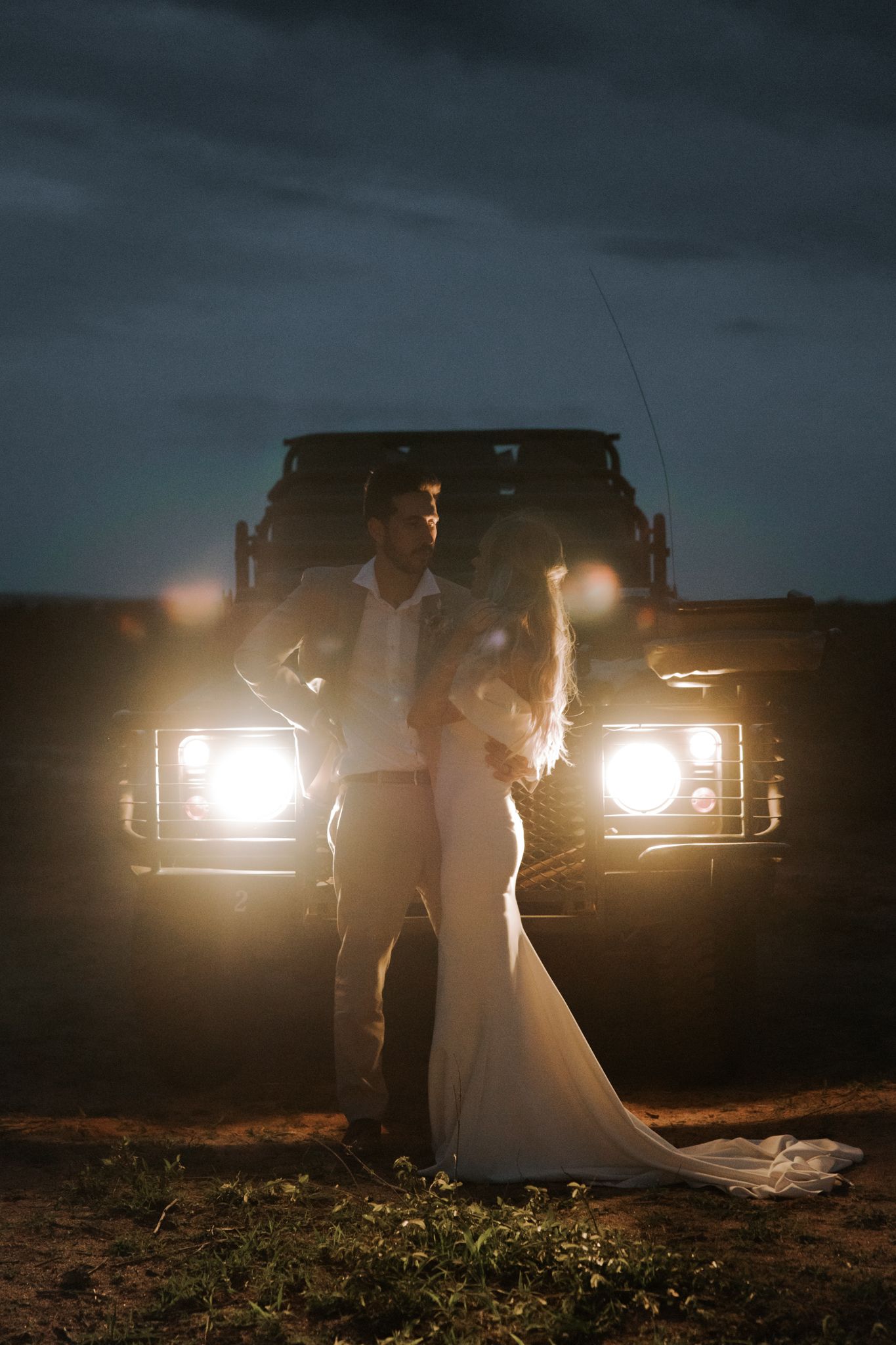 A silhouetted couple embraces in front of car headlights creating a romantic backlit scene at dusk.