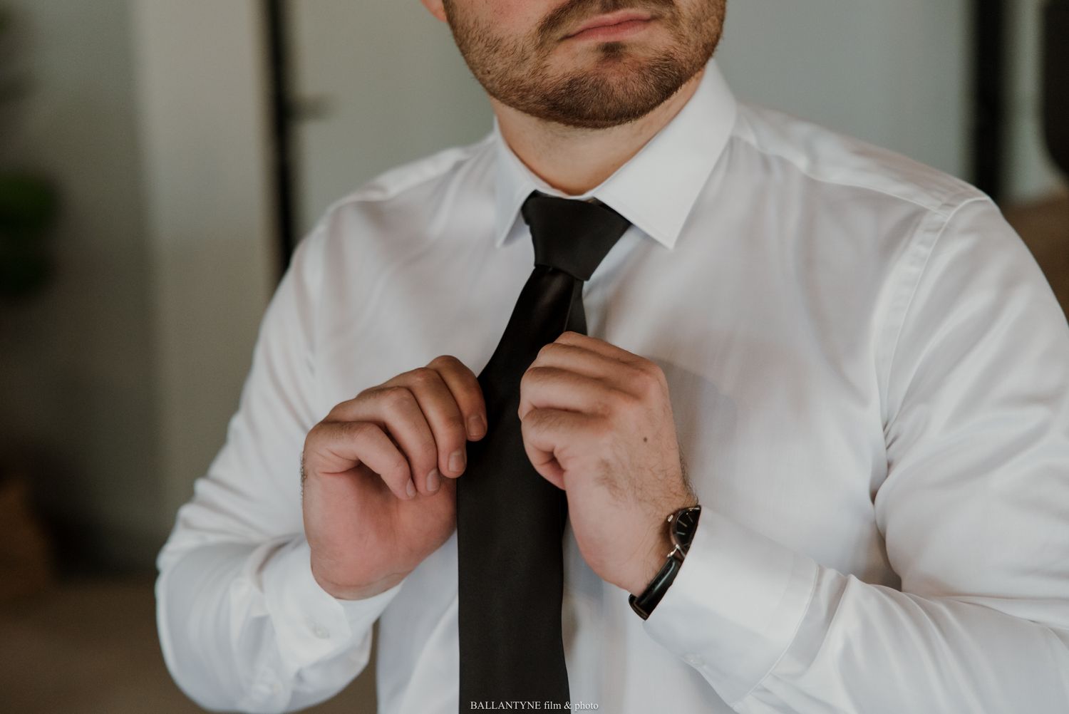 Man in white dress shirt adjusting black necktie, wearing a brown watch, getting dressed for formal occasion.