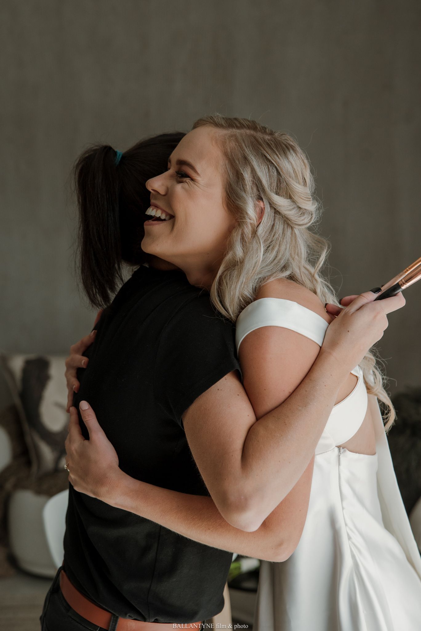 Two women share a joyful hug, one in a white off-shoulder dress, likely a bride, smiling warmly indoors.