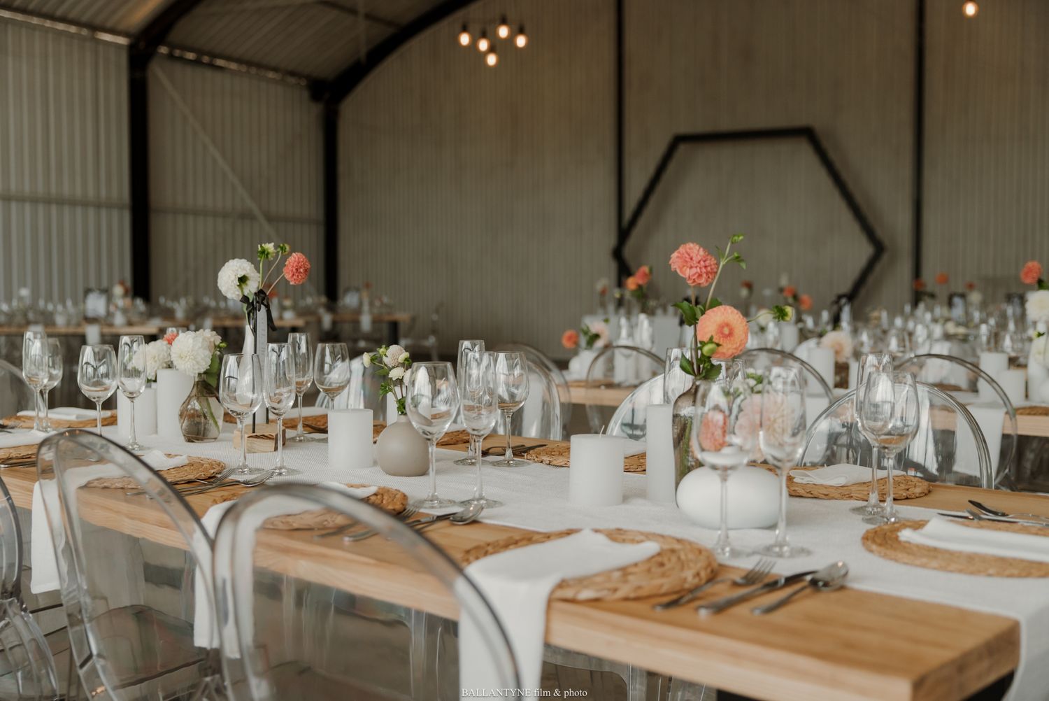 Elegant barn wedding reception with long wooden tables, white linens, ghost chairs, and pink floral centerpieces.