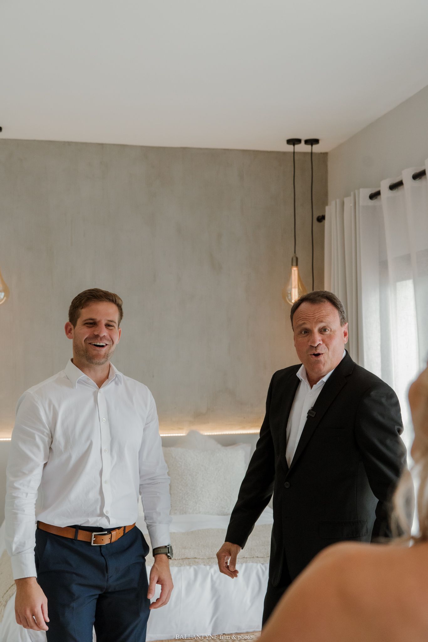 Two men smiling in a modern room, one in a white shirt and one in a black suit, standing near a white sofa.