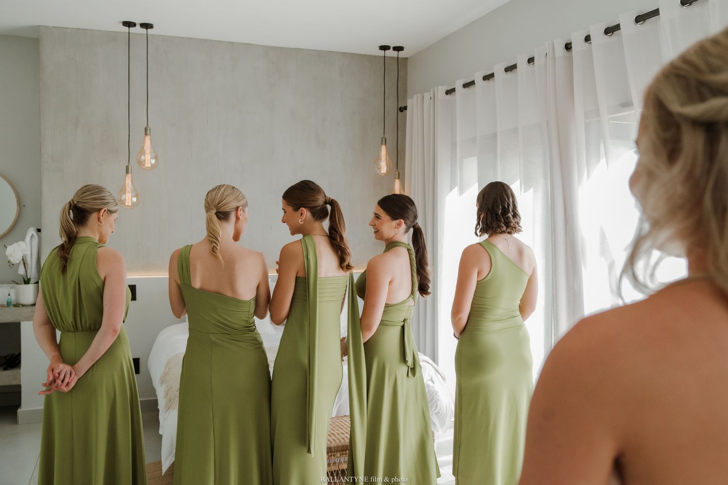 Bridesmaids in matching olive green dresses standing by a bright window during wedding preparations.