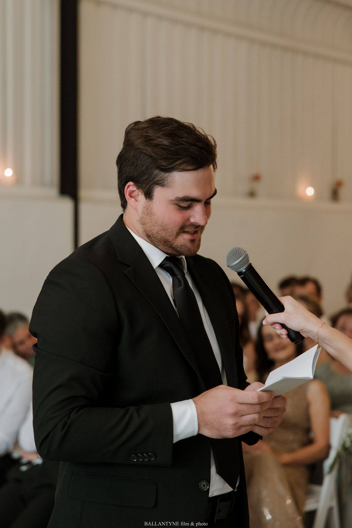 Man in black suit giving a wedding speech at a reception, holding notes and a microphone.