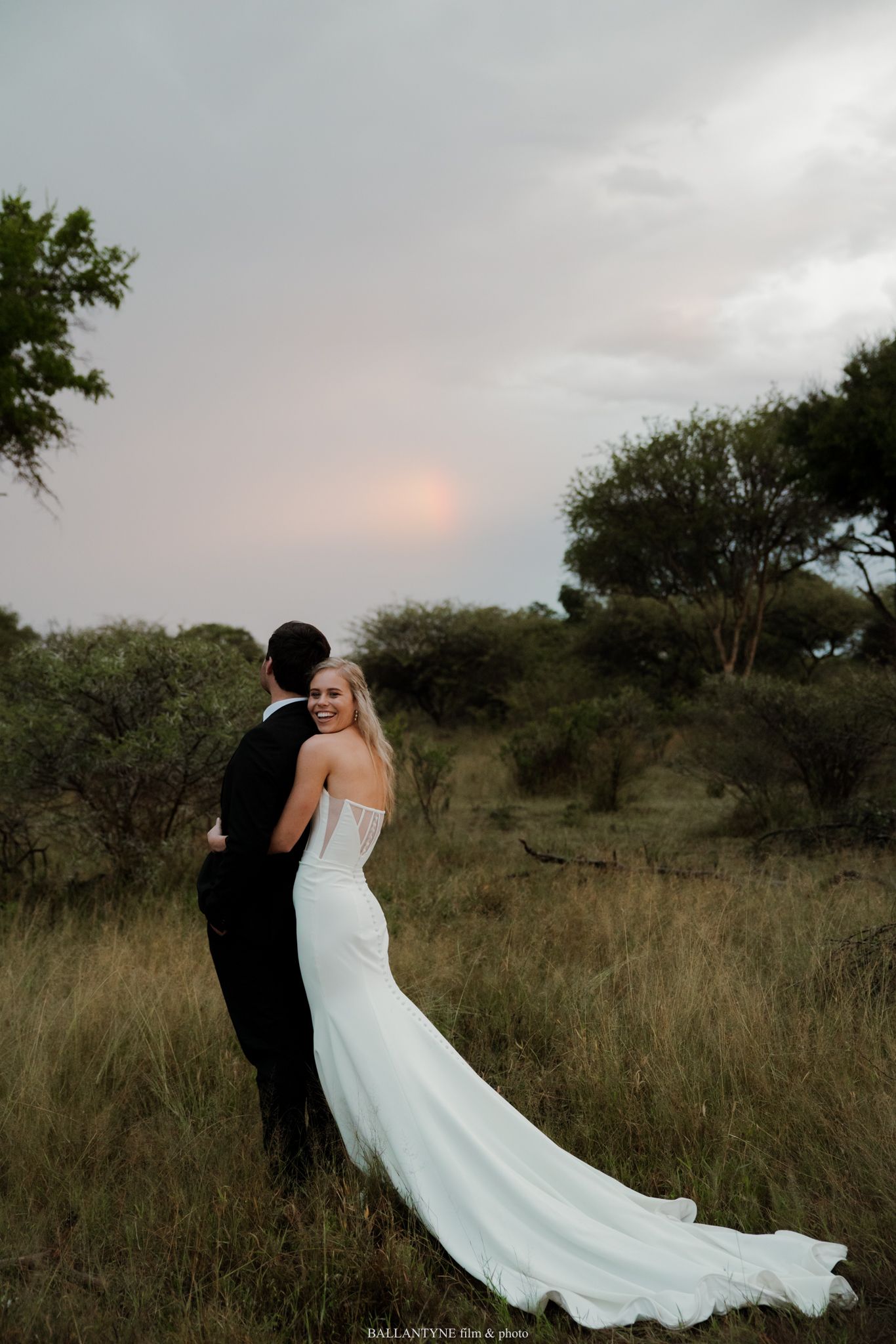 Bride in white gown with train smiles over shoulder as groom embraces her in lush African savanna at sunset.