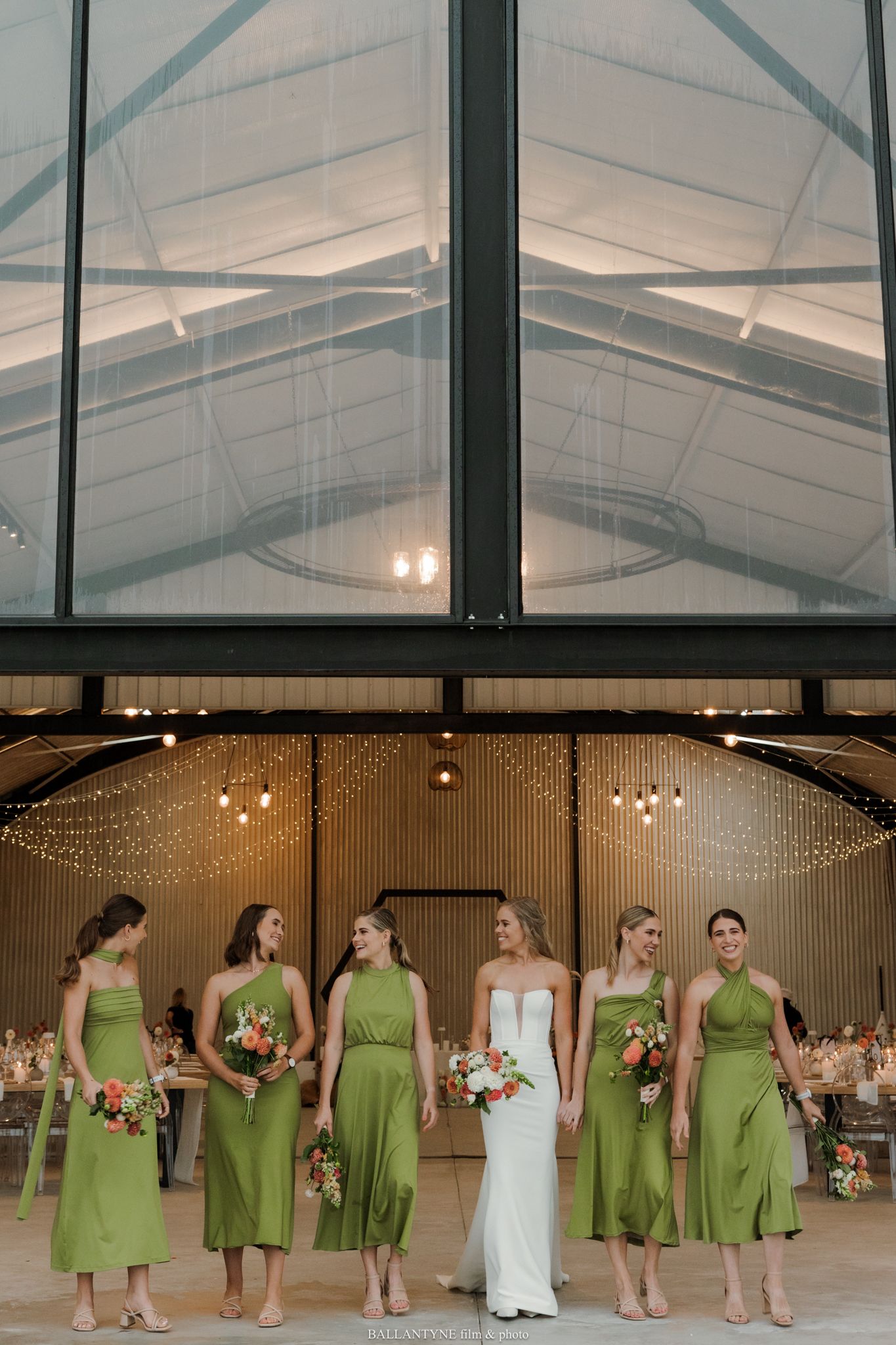 Bride in white gown stands with five bridesmaids in green midi dresses inside an elegant barn wedding venue.