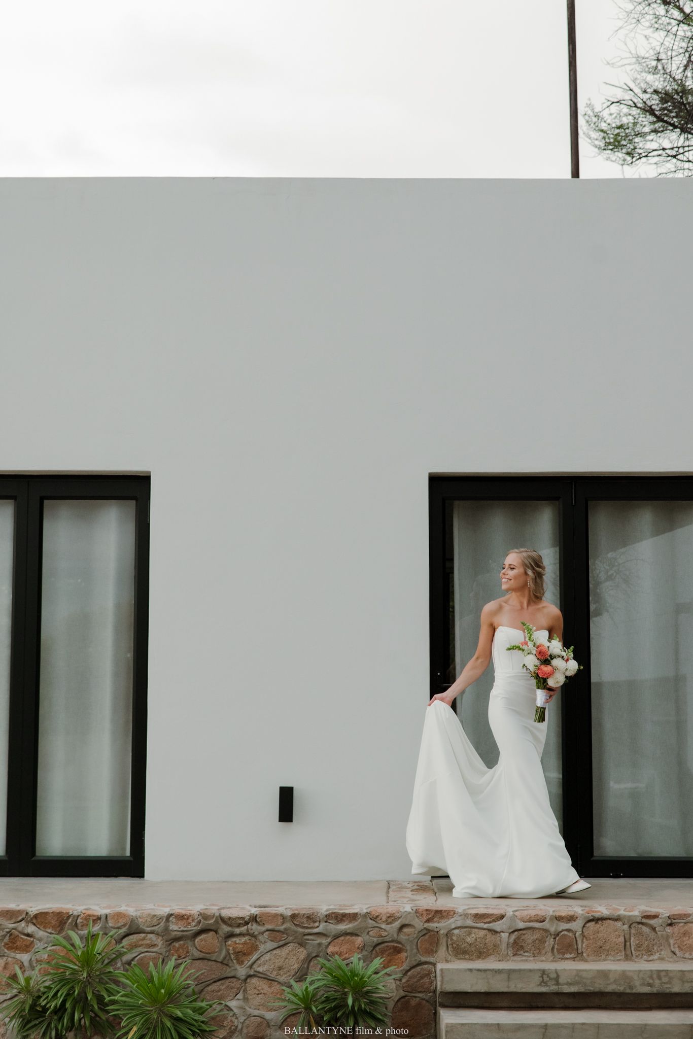 Bride in white strapless gown holding bouquet stands by modern white building with black-framed windows.
