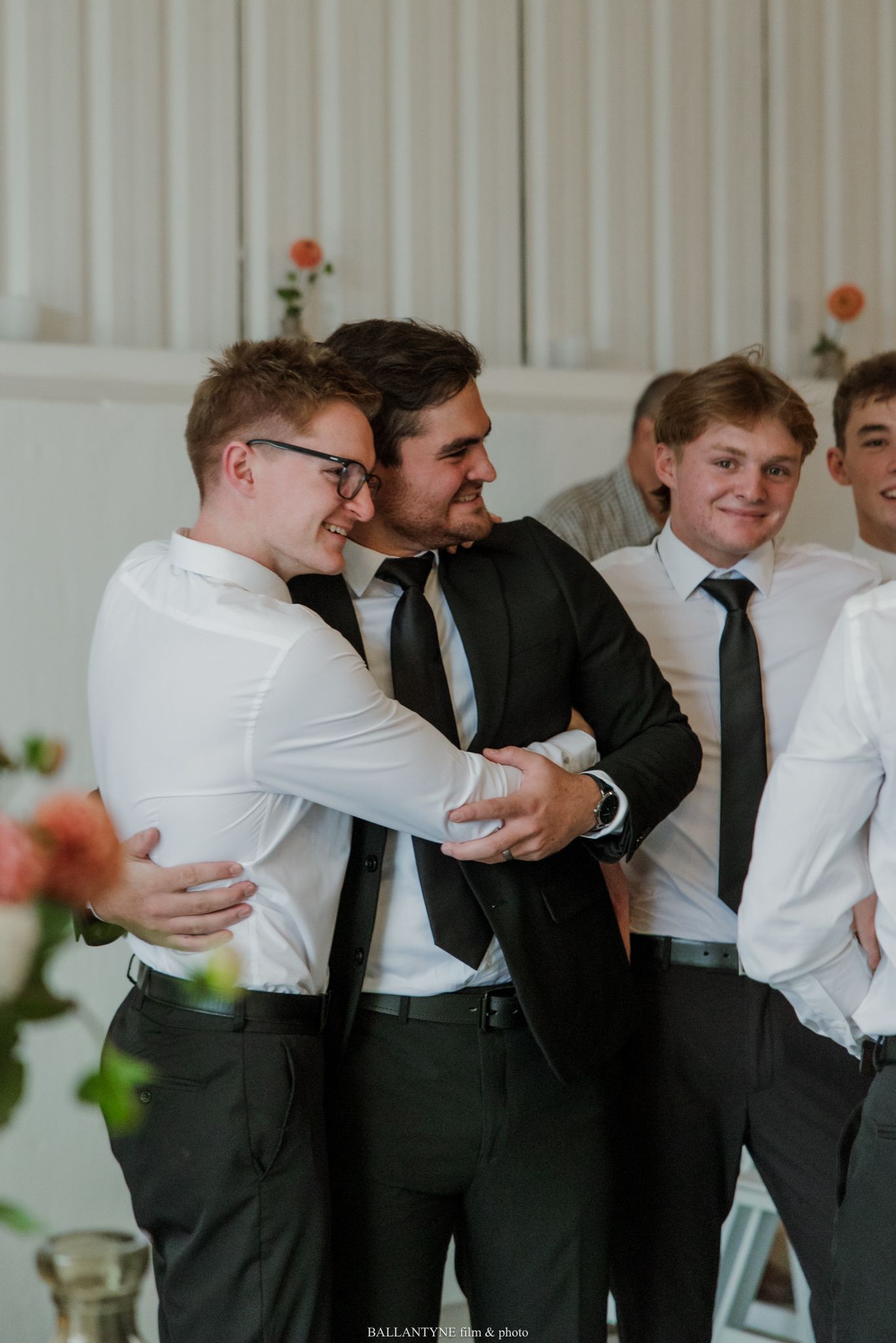 Young men in formal attire sharing a joyful hug at a wedding reception event.
