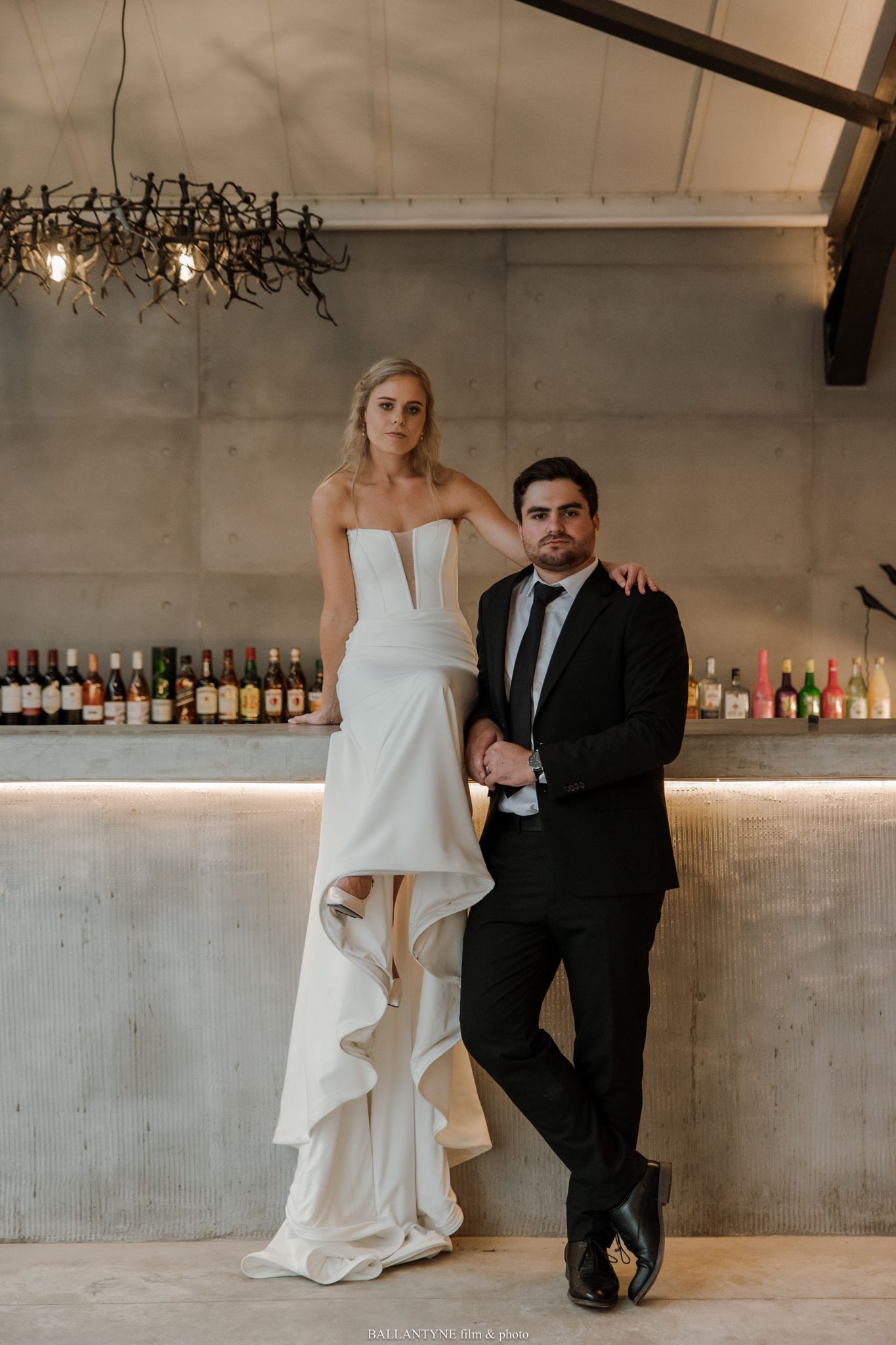Bride in strapless white gown with ruffles stands with groom in black suit at elegant bar venue.